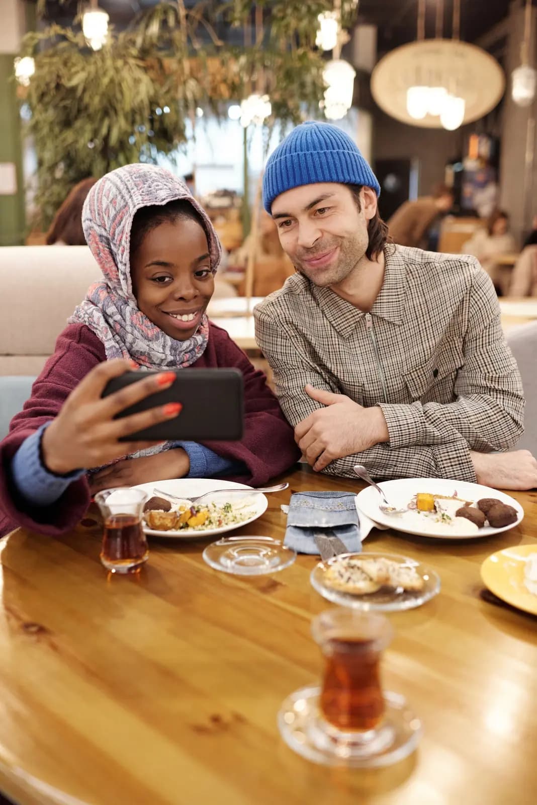 Two friends at a food hall table laugh at a phone one of them holds up, with plates of food and glasses of tea in front of them.