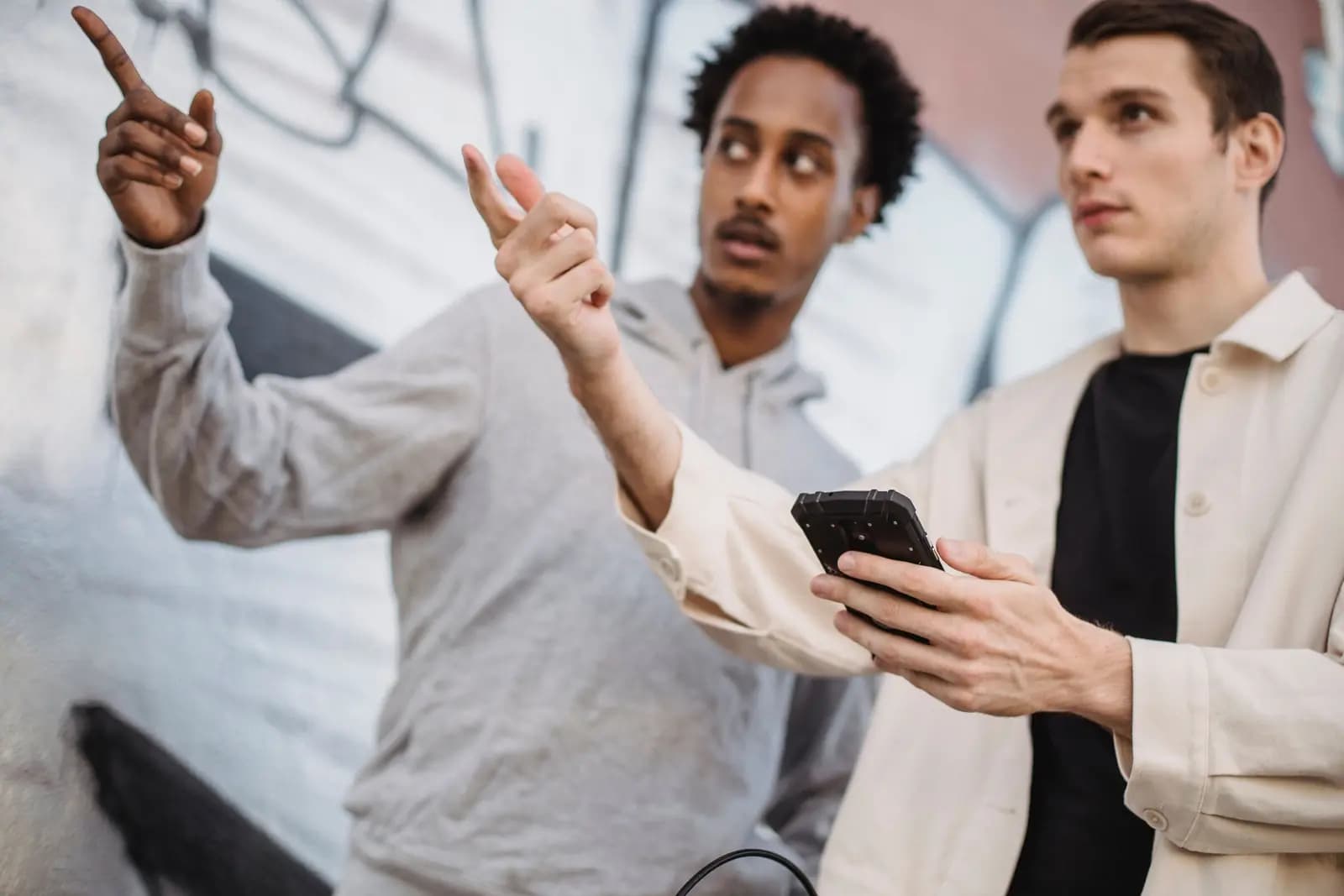 Two friends point up in front of a painted mural on a city wall, one of them holding a phone.