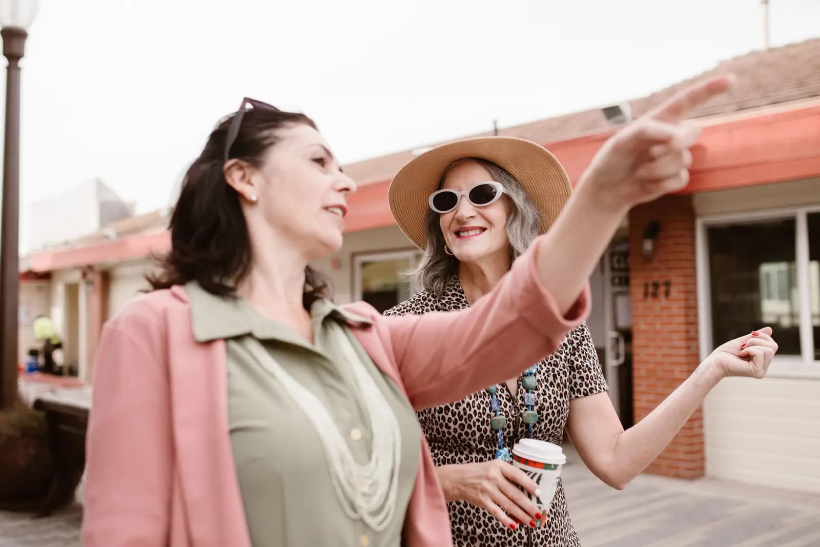 Two women on a small-town main street, one pointing up the street while the other holds a coffee cup.
