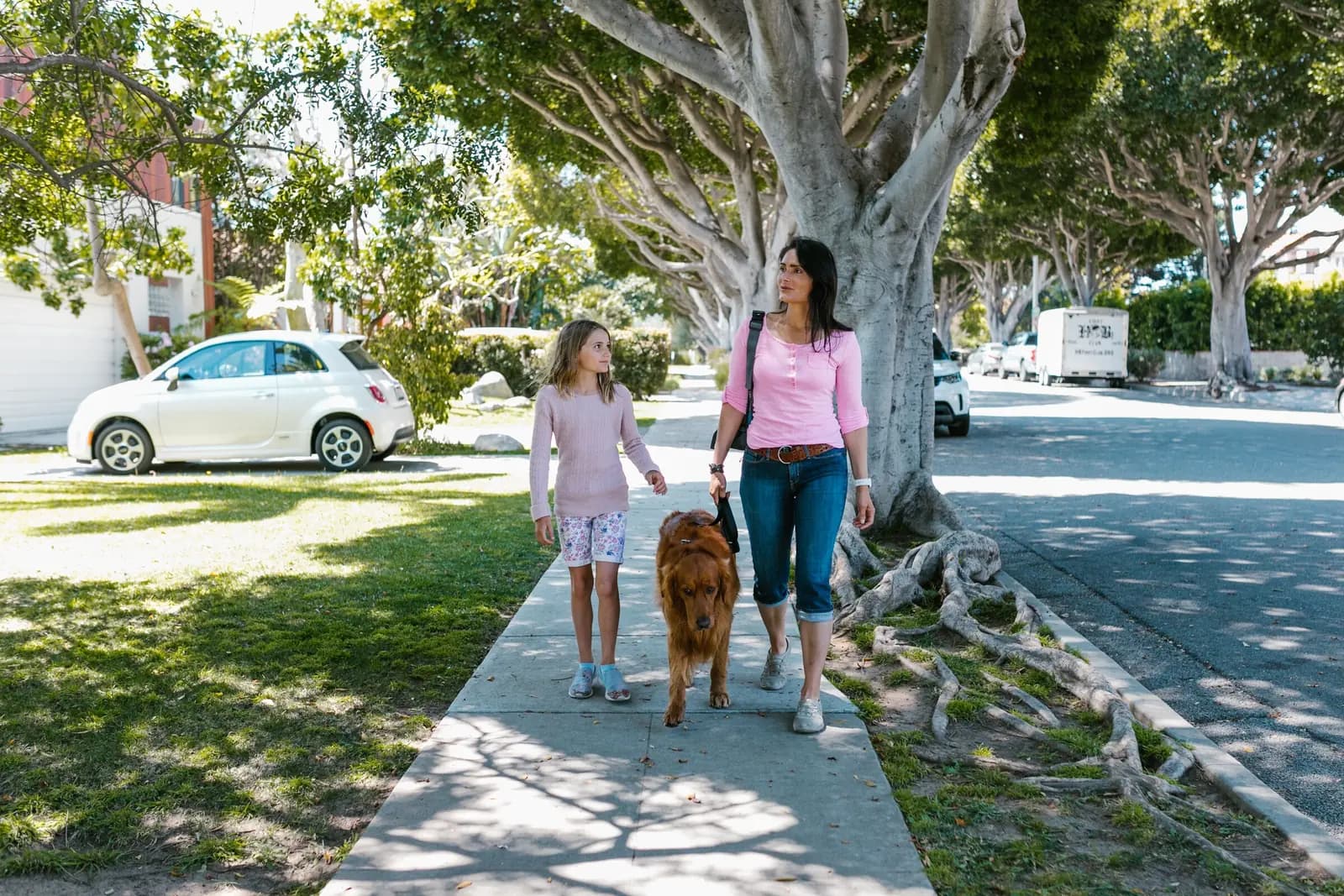 A mother and her daughter walk a golden retriever down a sunny residential sidewalk.