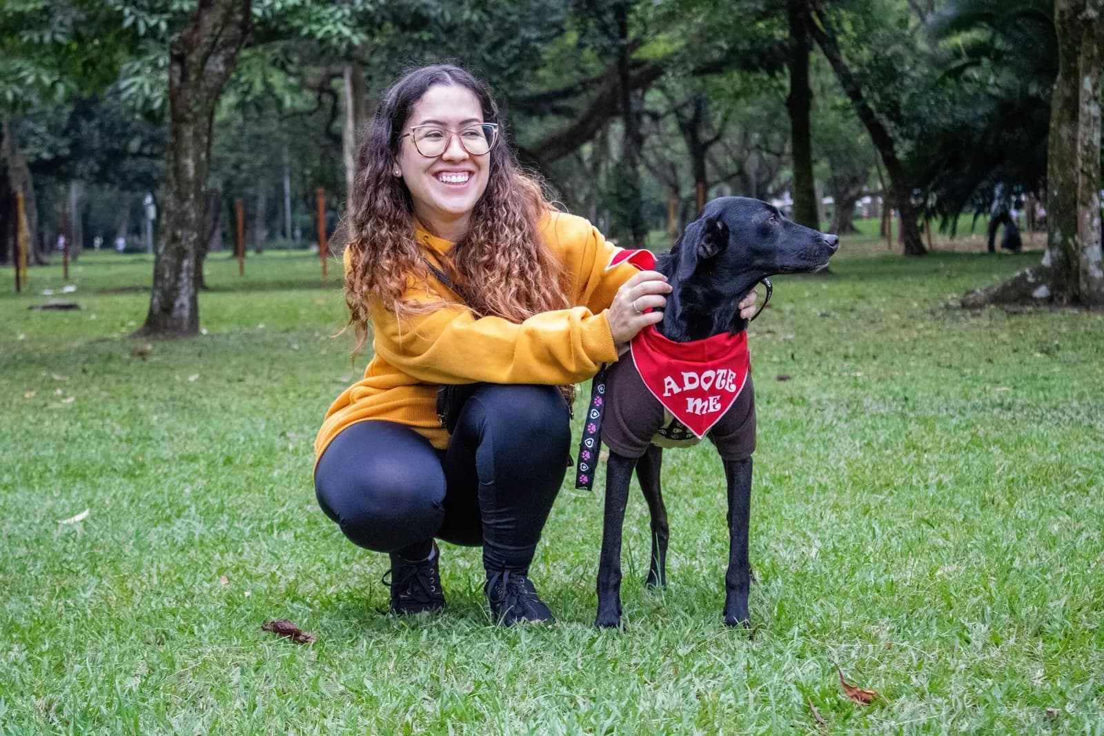 A smiling woman crouches in a park beside a black dog wearing a red Adopt me bandana.