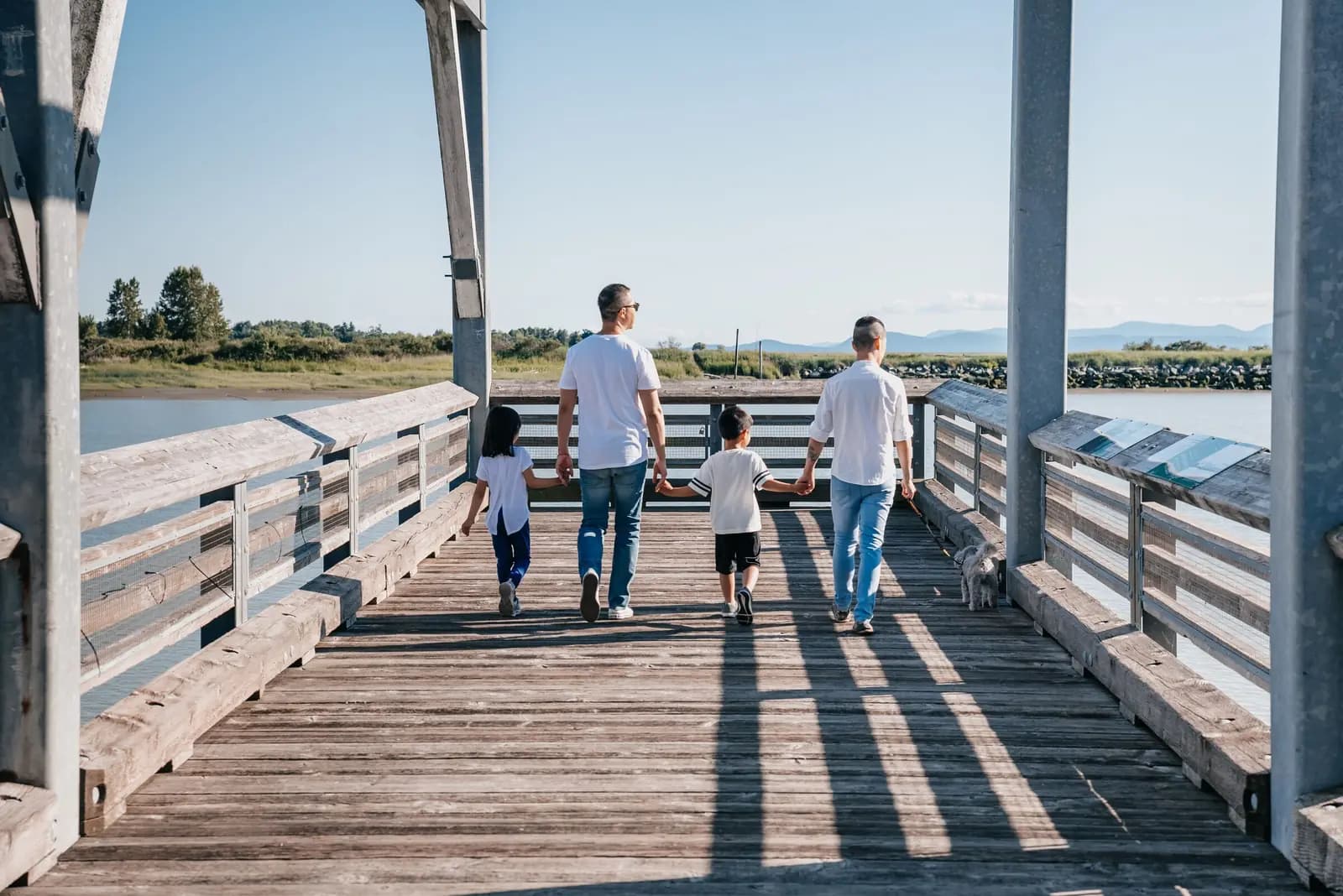 A family of four walks hand in hand along a wooden pier toward the water on a sunny day.