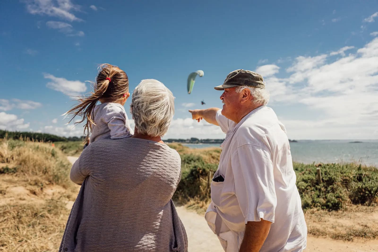 A grandfather points out to sea while a grandmother holds a small child beside him on a coastal path.