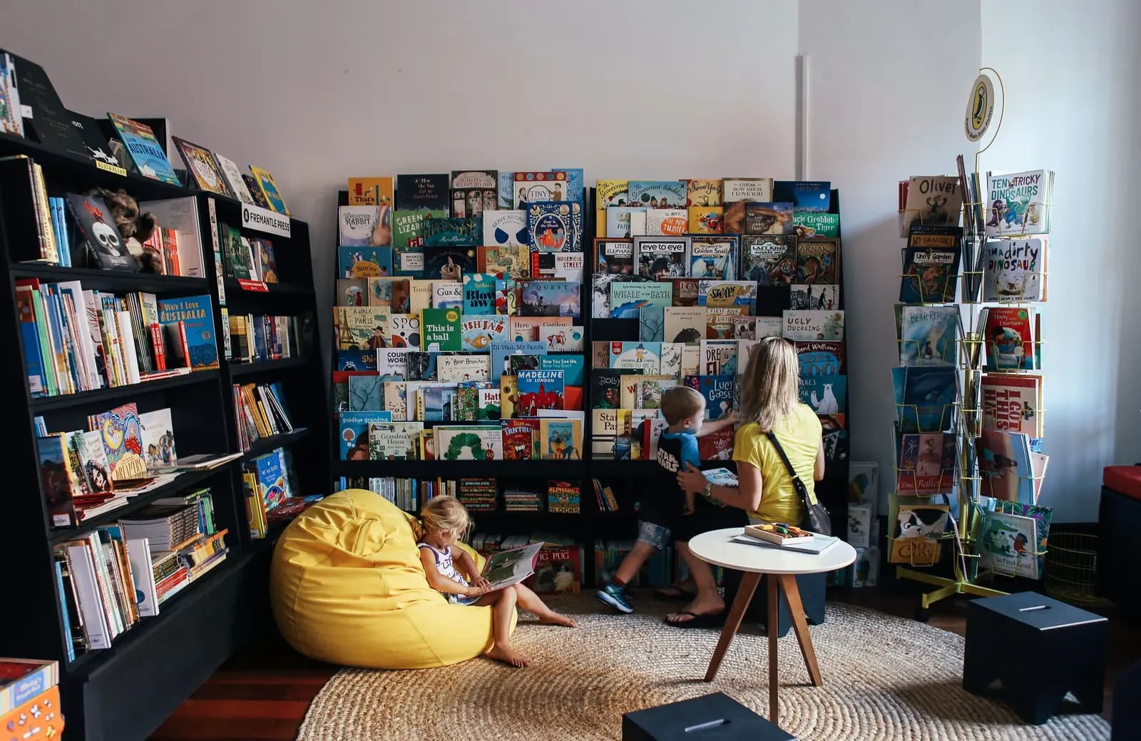 A bookstore children's corner with picture-book racks, a yellow beanbag and a parent browsing with two children.