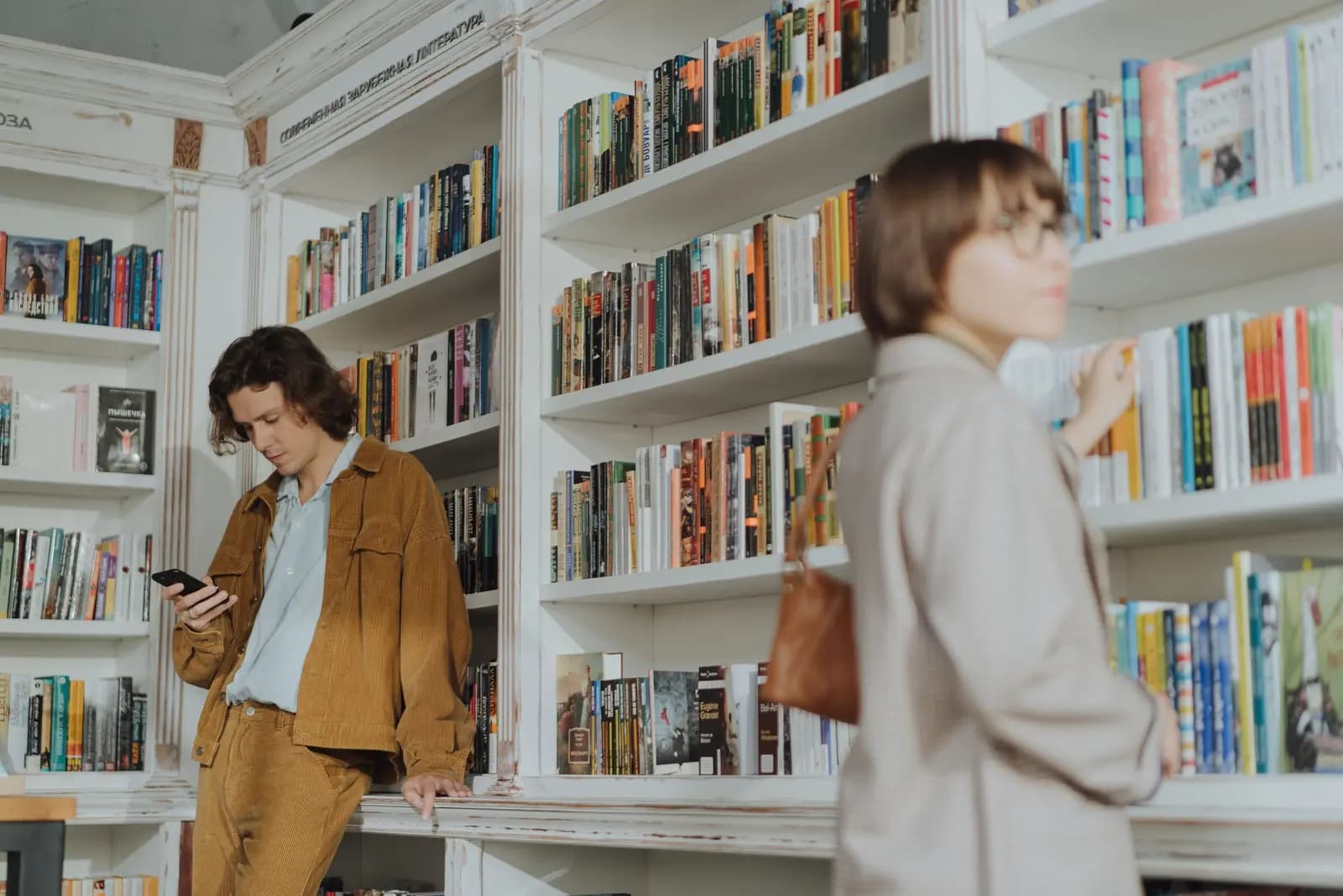 Two friends stand between tall white bookshelves in a bookstore, one checking a phone while the other reaches for a book.