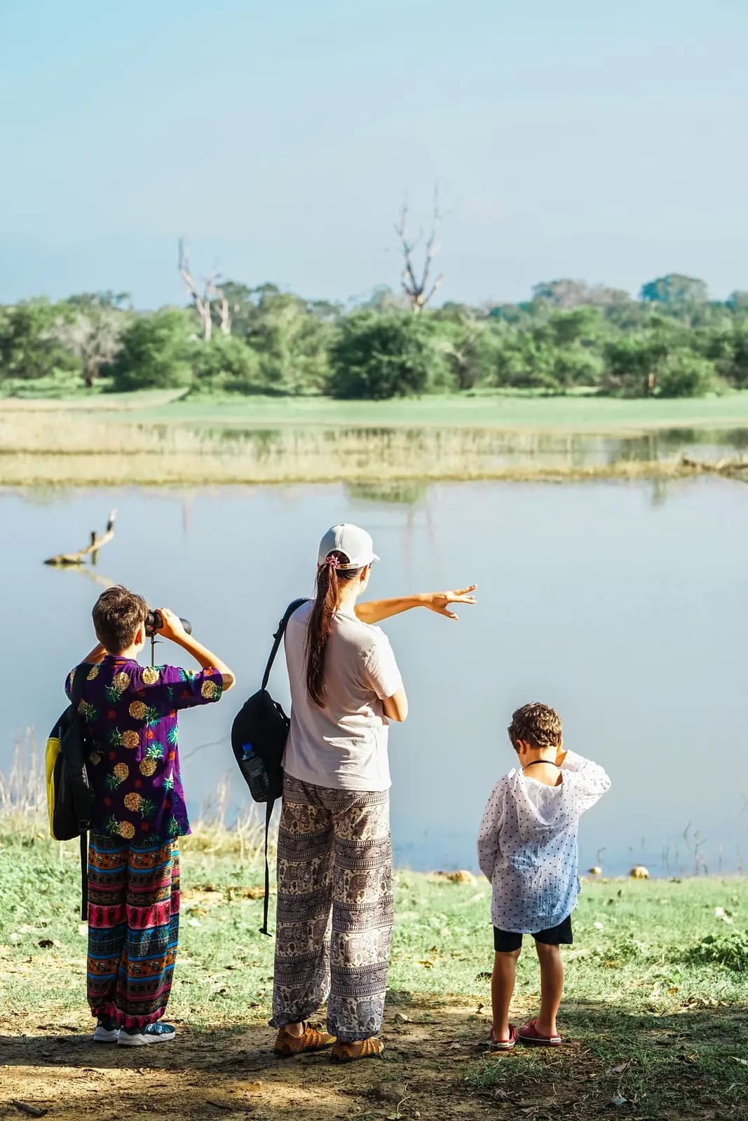 A mother points across a lake while one child looks through binoculars and another stands at the shore, seen from behind.