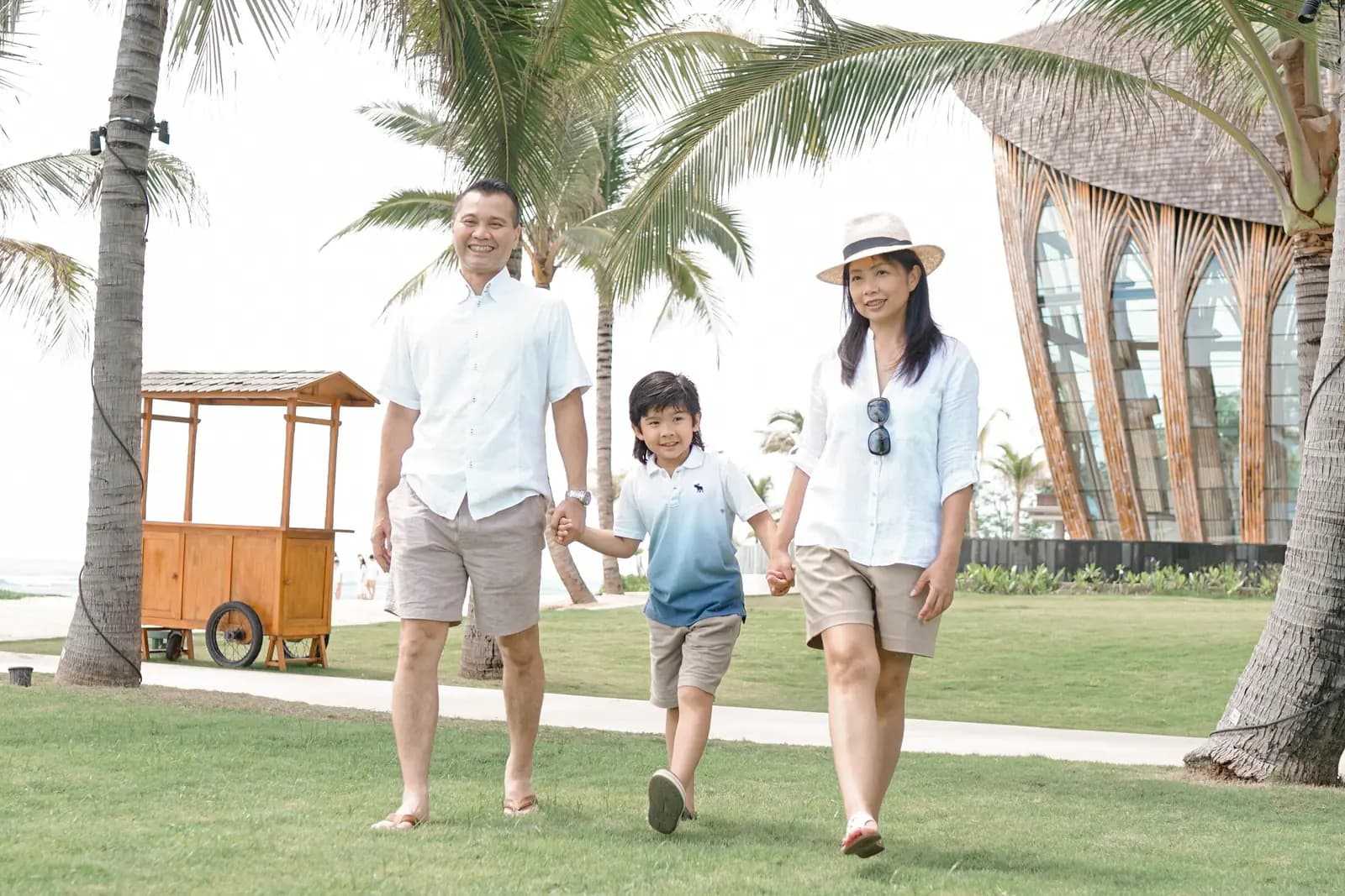 A family of three walk hand in hand across a resort lawn under palm trees.