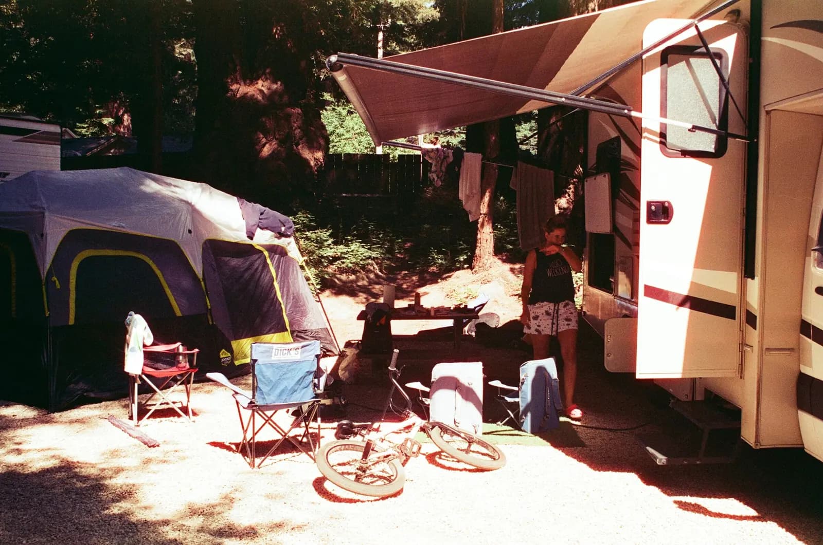 A campsite in the woods with a tent, camp chairs and a bike beside an RV with its awning out.