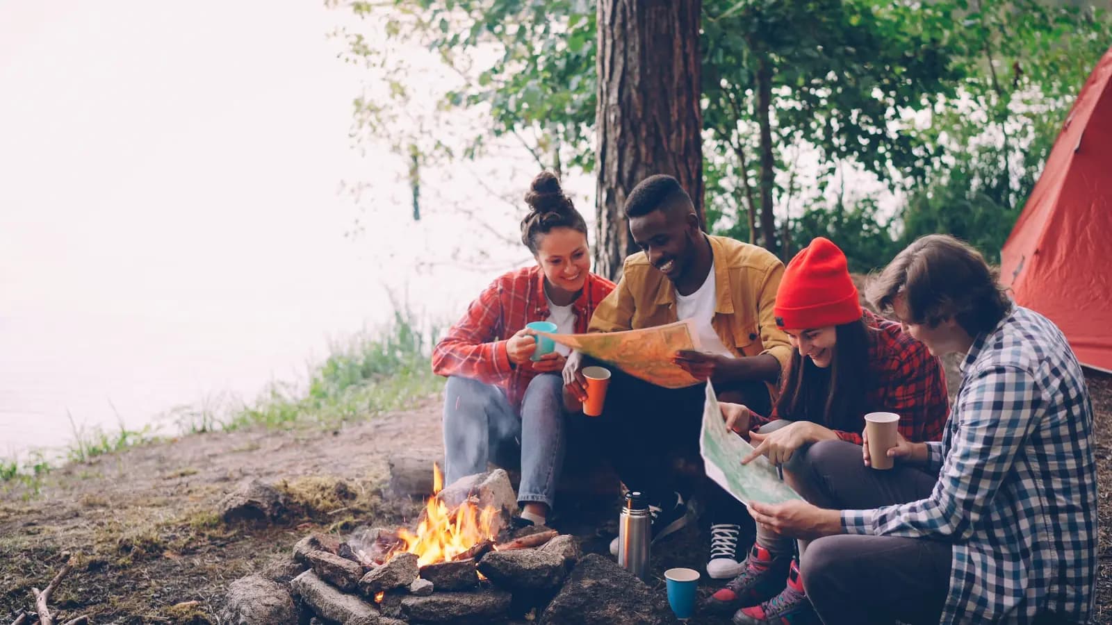 Four friends sit around a campfire beside a lake with camp mugs and a paper map.