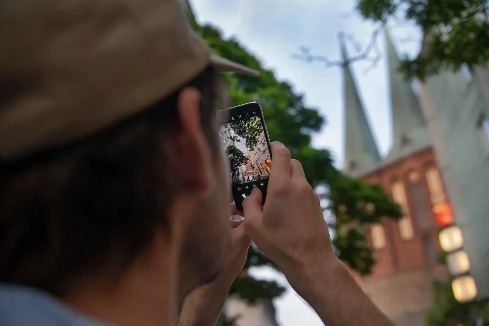 A man in a cap holds up a phone to photograph a church's twin spires.
