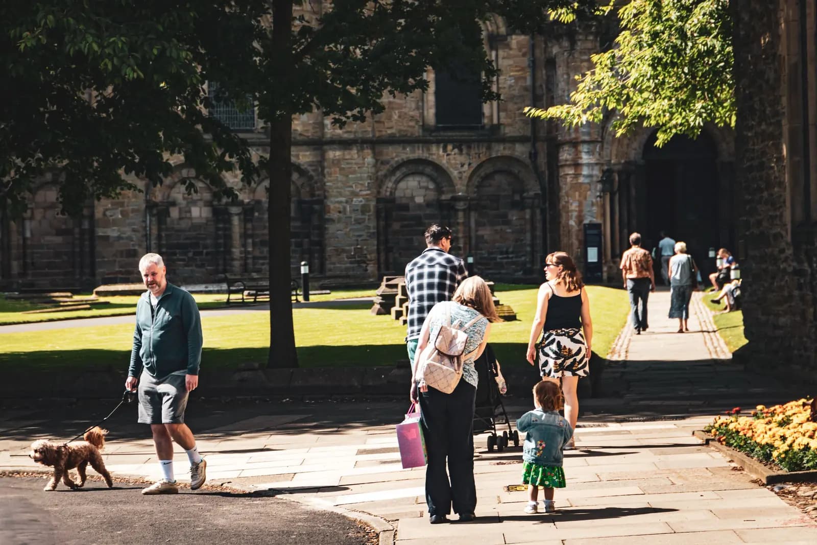 People cross a sunny lawn and path away from an old stone church, a toddler walking with her mother and a man with a dog.