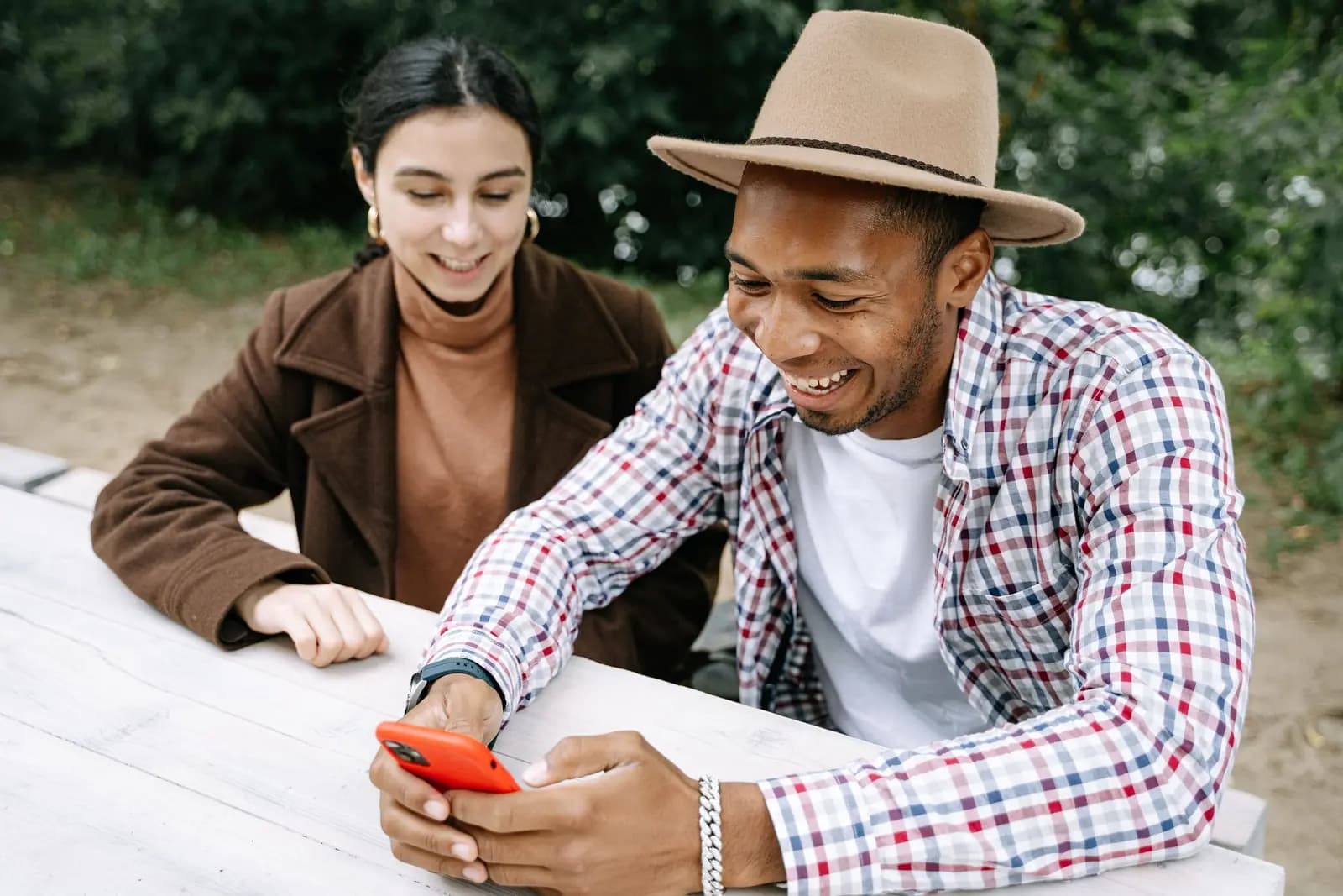 Two adults laugh together over a phone at a wooden picnic table outdoors.
