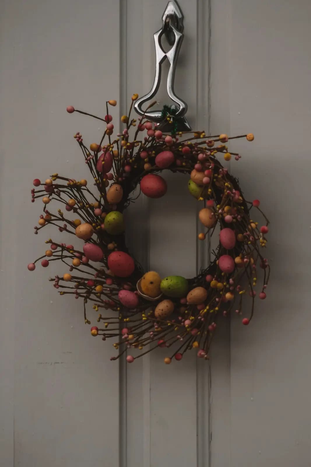 A twig wreath studded with speckled pastel eggs hanging on a grey front door.