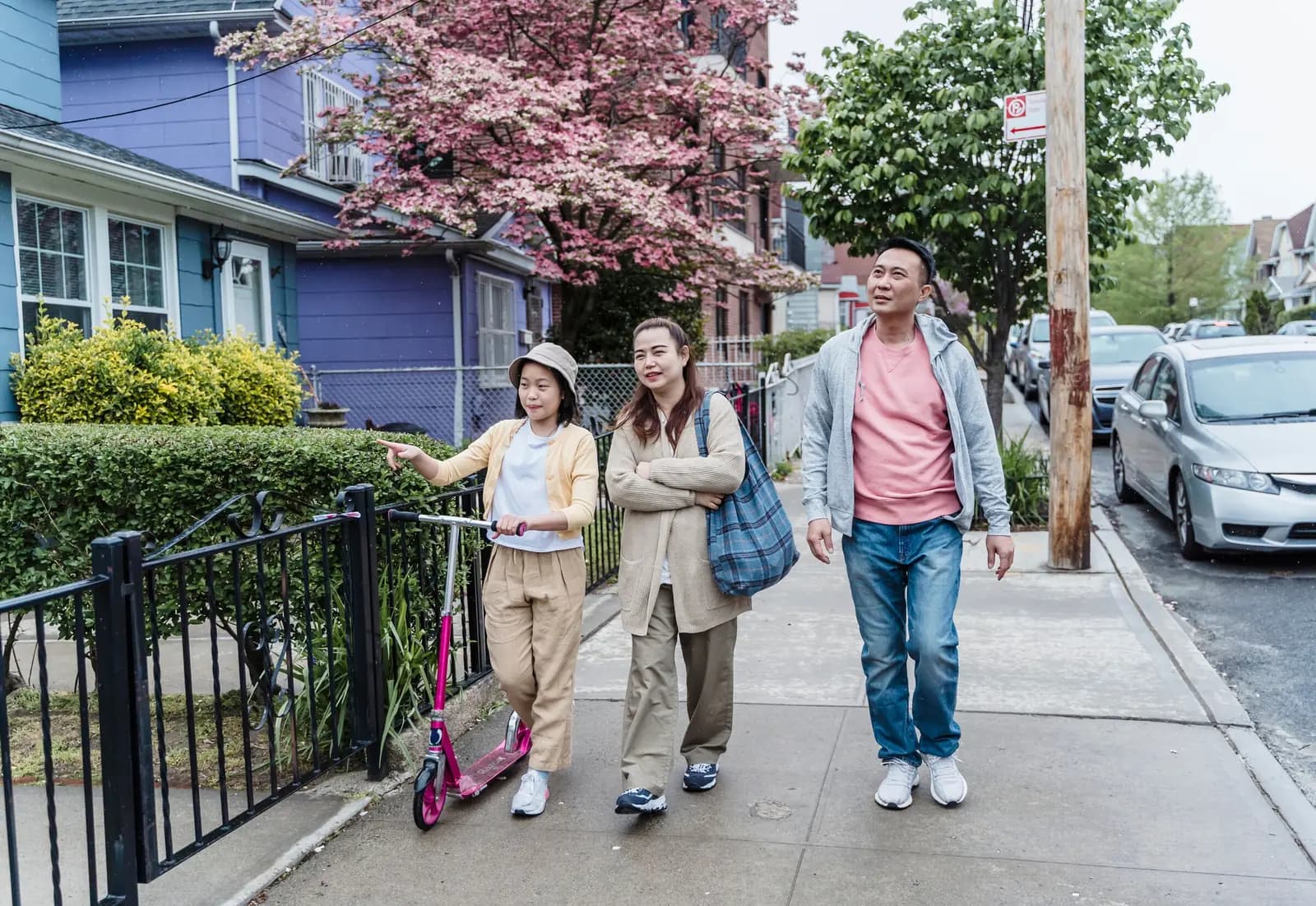 A family walks along a spring sidewalk past a blossoming tree and painted houses, a girl with a scooter pointing ahead.