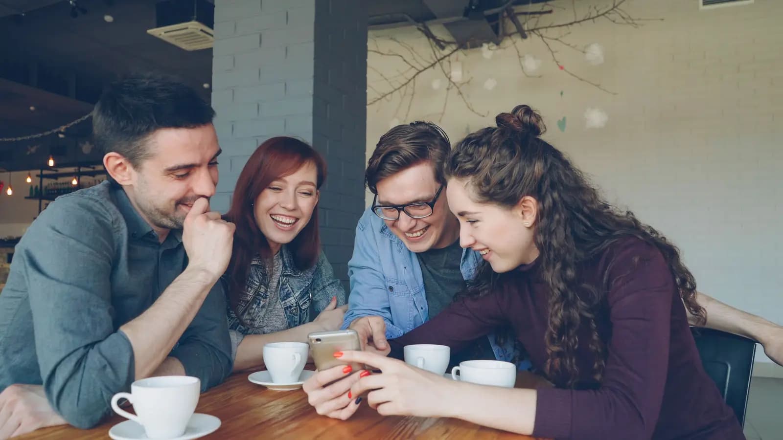 Four friends lean in together over a phone at a cafe table with coffee cups.
