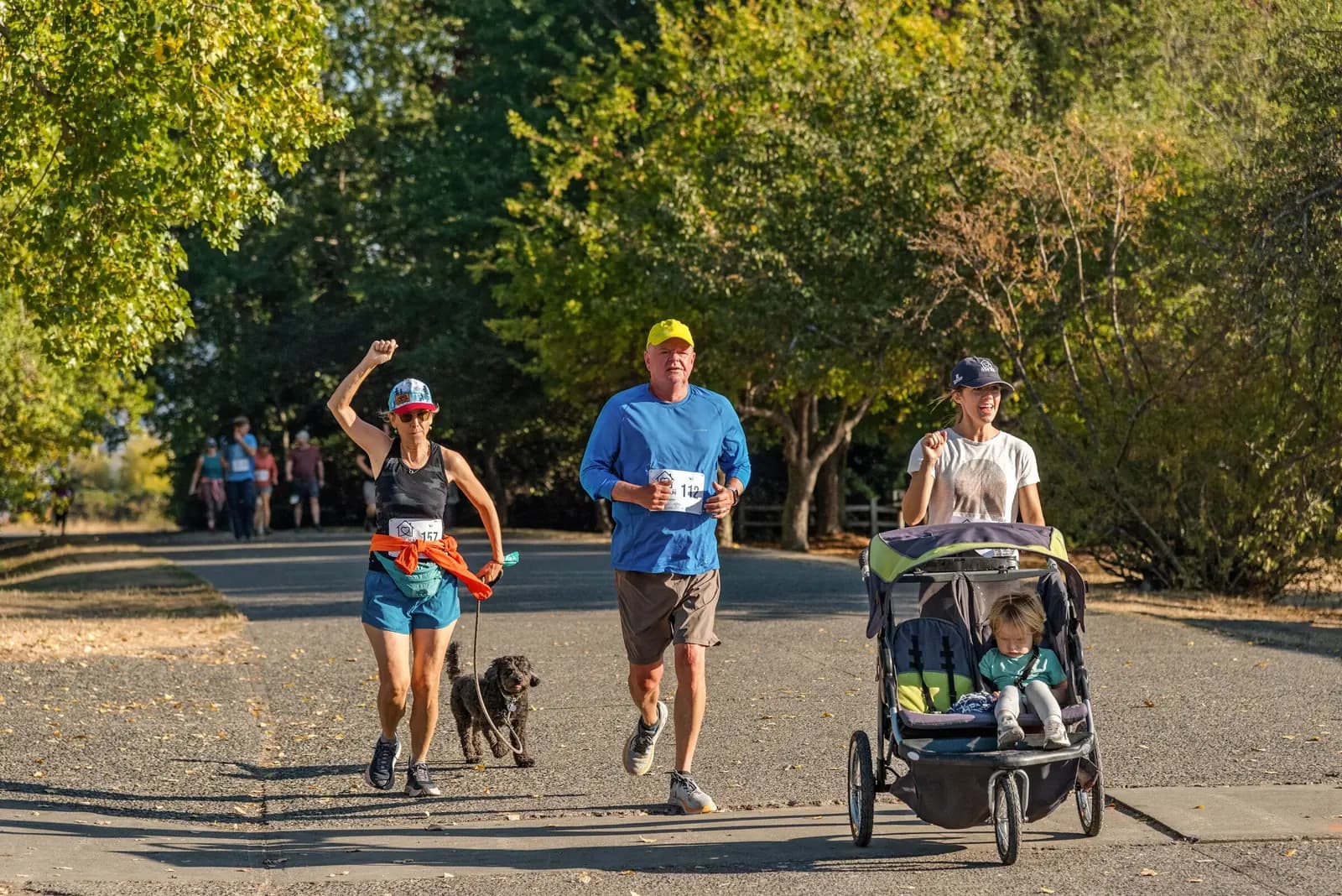 Runners in race bibs on a tree-lined path, one with a small dog on a leash and one pushing a running stroller.