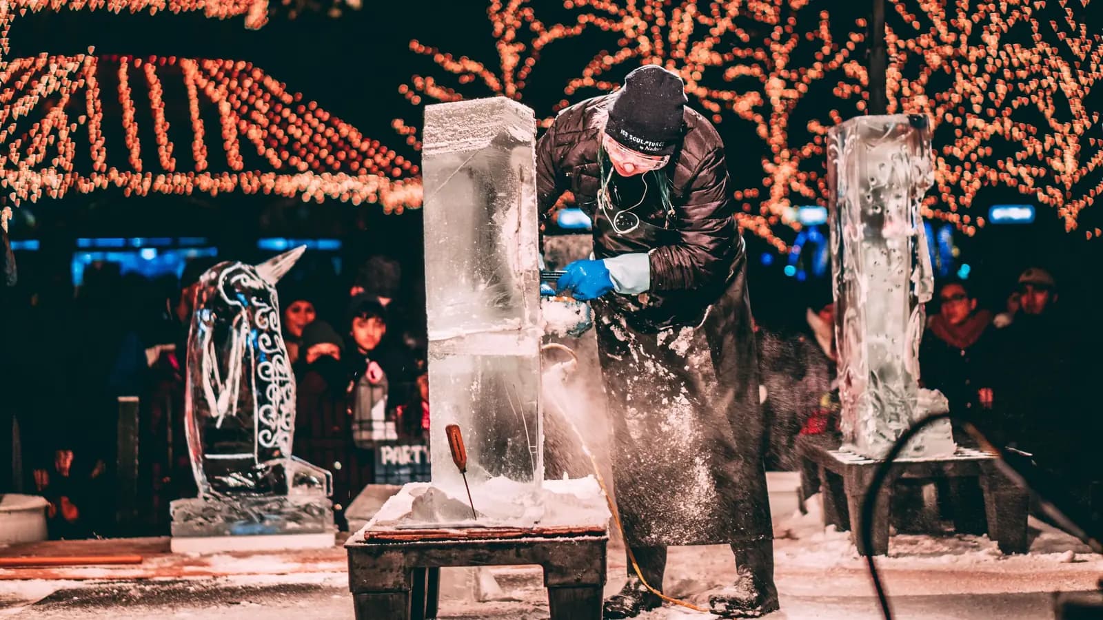 An ice sculptor works at night beside finished sculptures, with lights strung overhead.