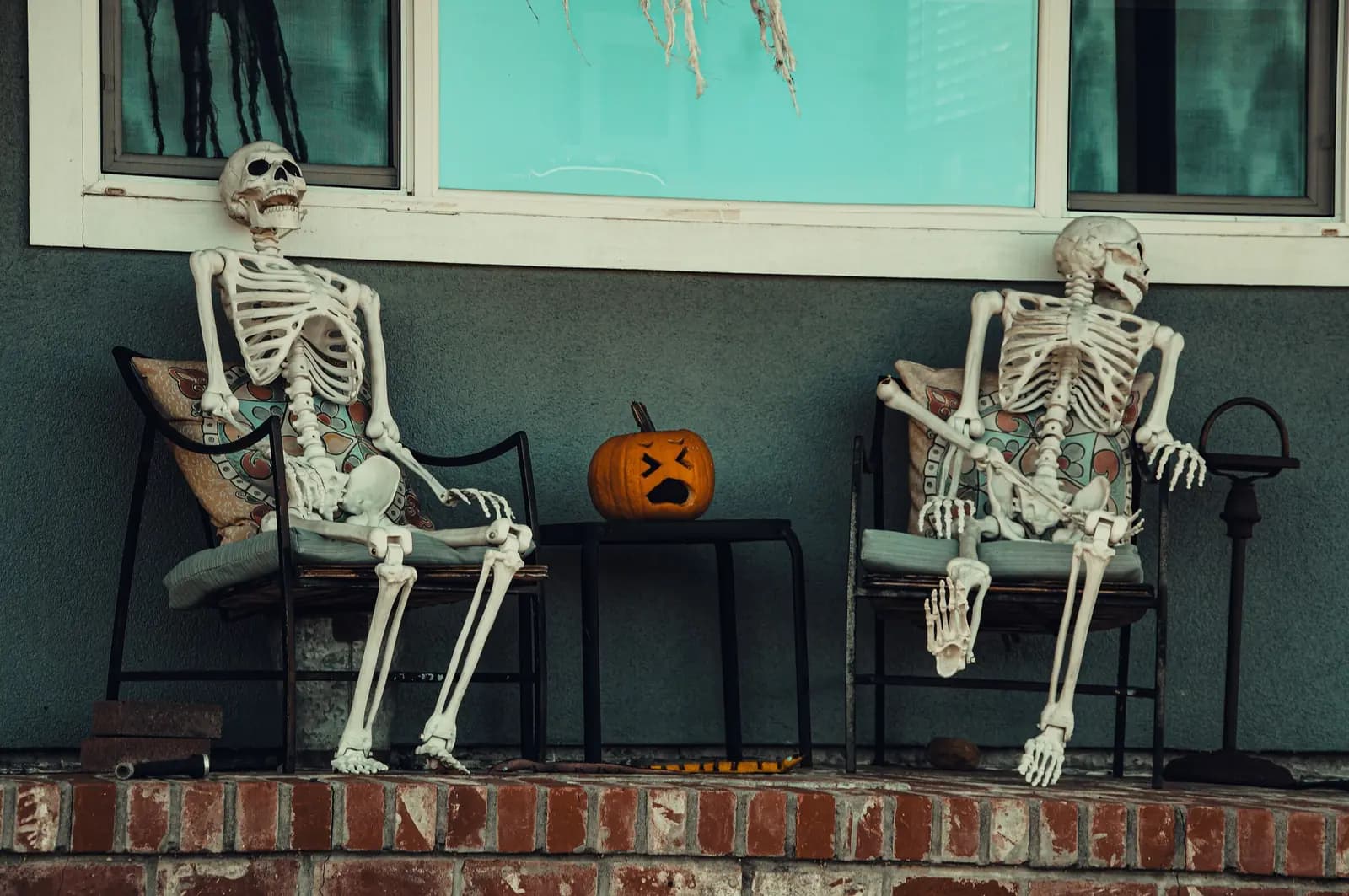 Two skeletons sitting in chairs beside a carved pumpkin on a decorated Halloween porch.