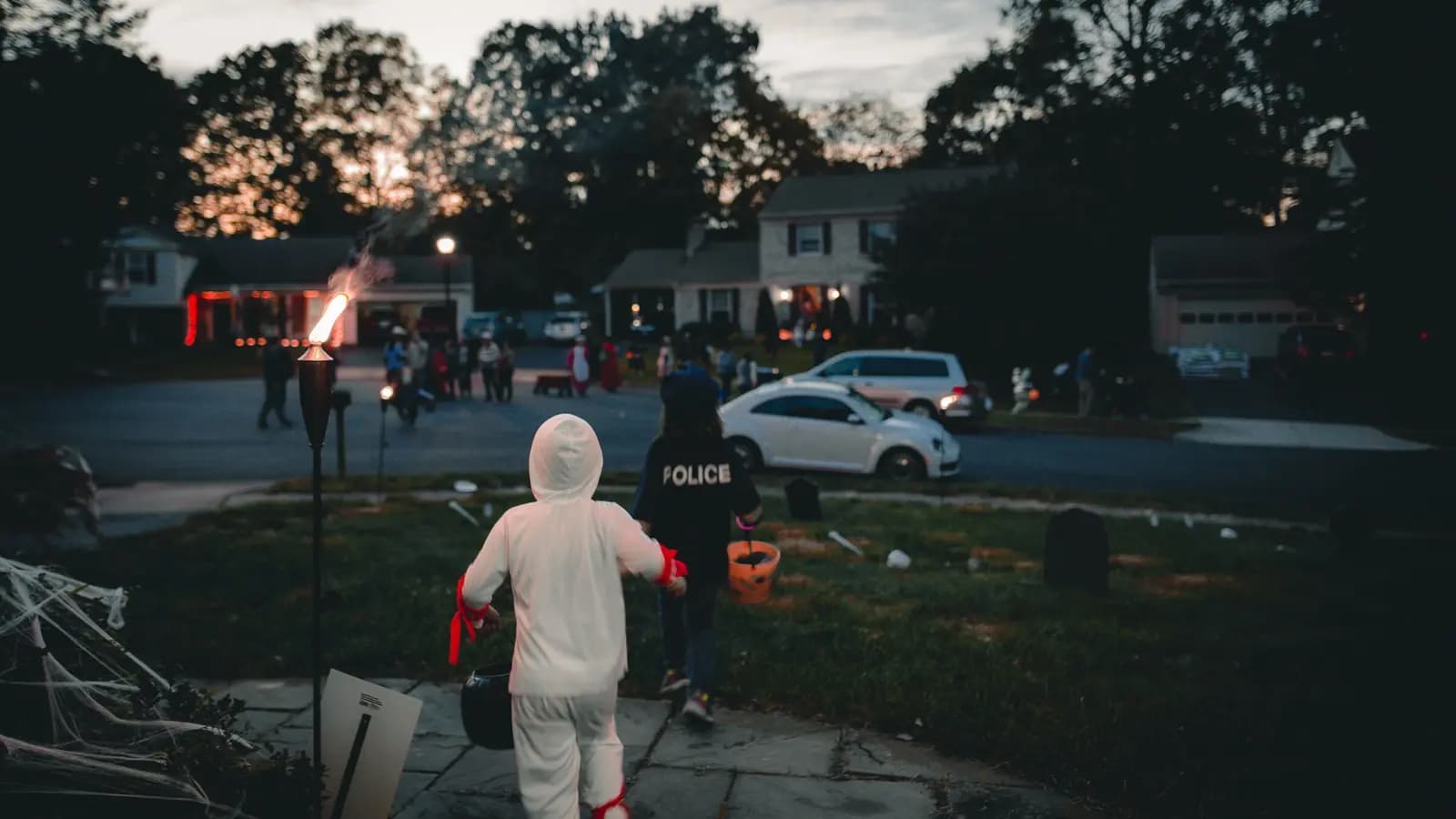 Two children in costumes walk past a lit torch and tombstones toward a street of houses at dusk on Halloween.