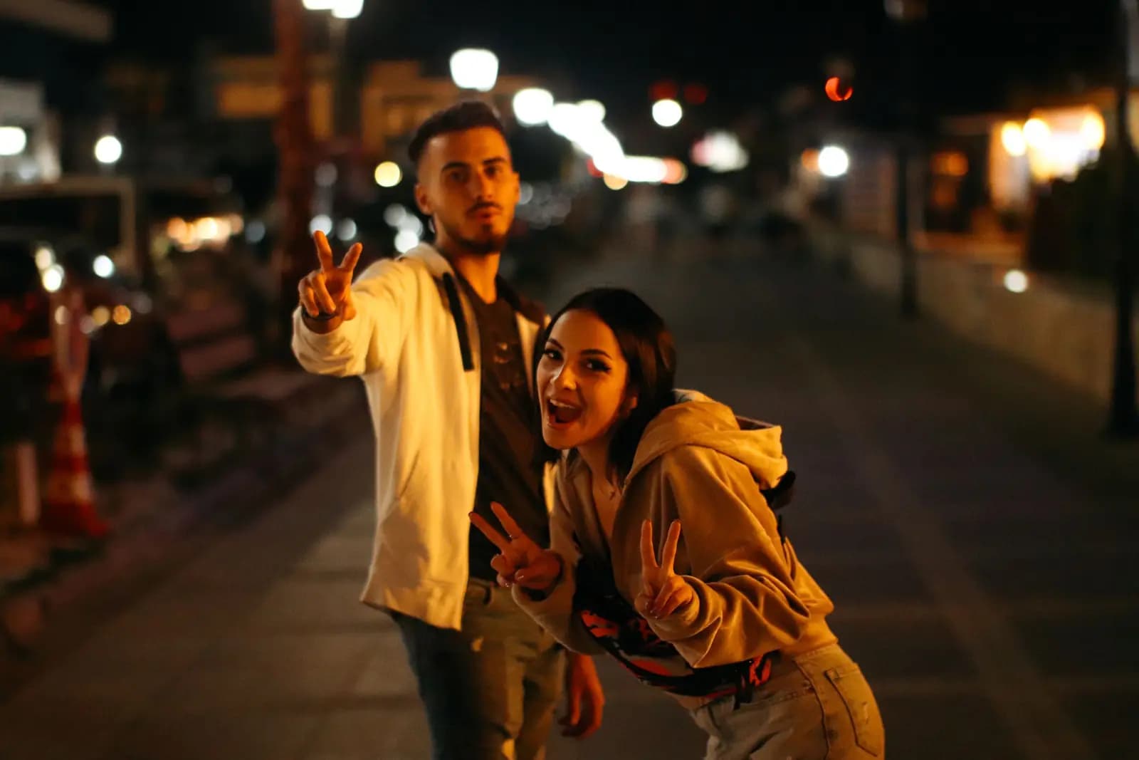 Two young friends pull playful faces and make peace signs on a lit street at night.