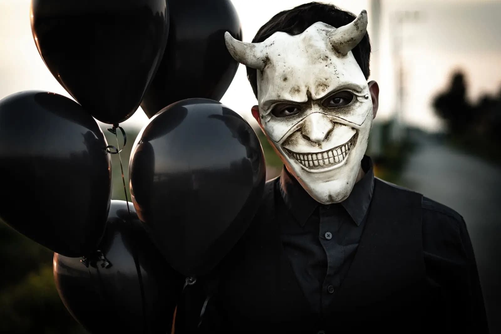 A person in a grinning horned white mask holds a bunch of black balloons outdoors.