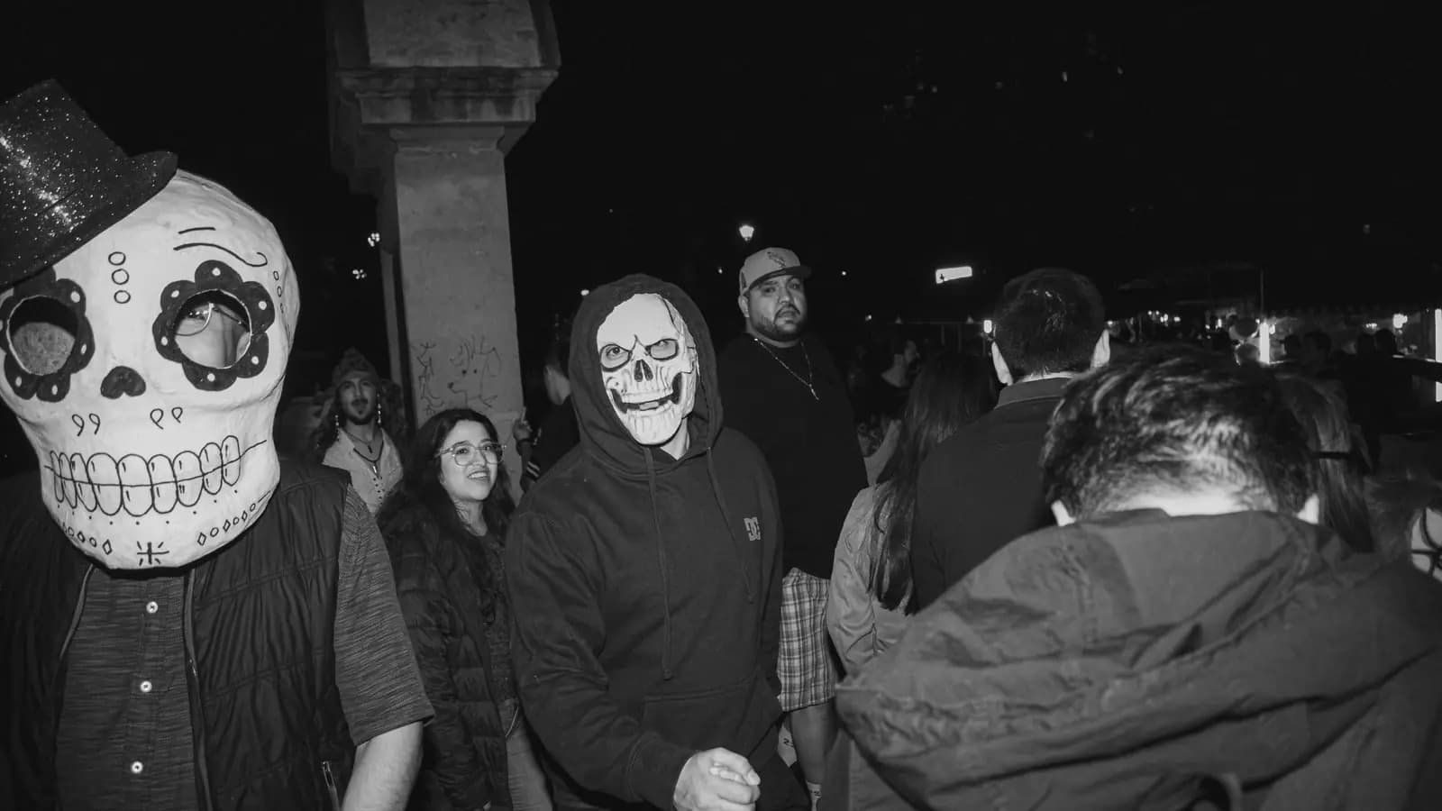 A black and white photo of a nighttime Halloween crowd, with two people in skull masks at the front.