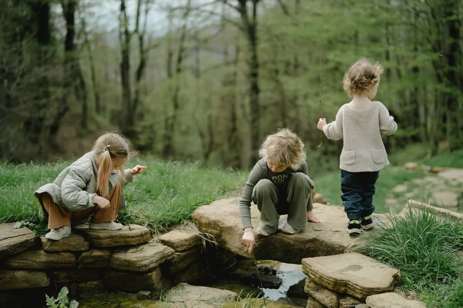 Three children of different ages crouch and stand on flat stones beside a small stream, looking into the water.