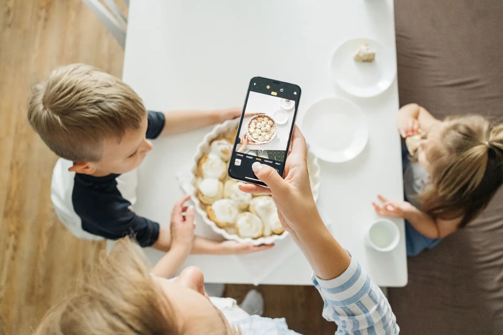 A parent holds a phone over a kitchen table while two children lean in around a dish, seen from above.