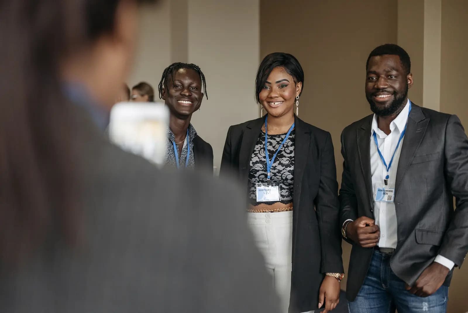 Three attendees with lanyards smile in a bright event room while someone in the foreground holds up a phone.