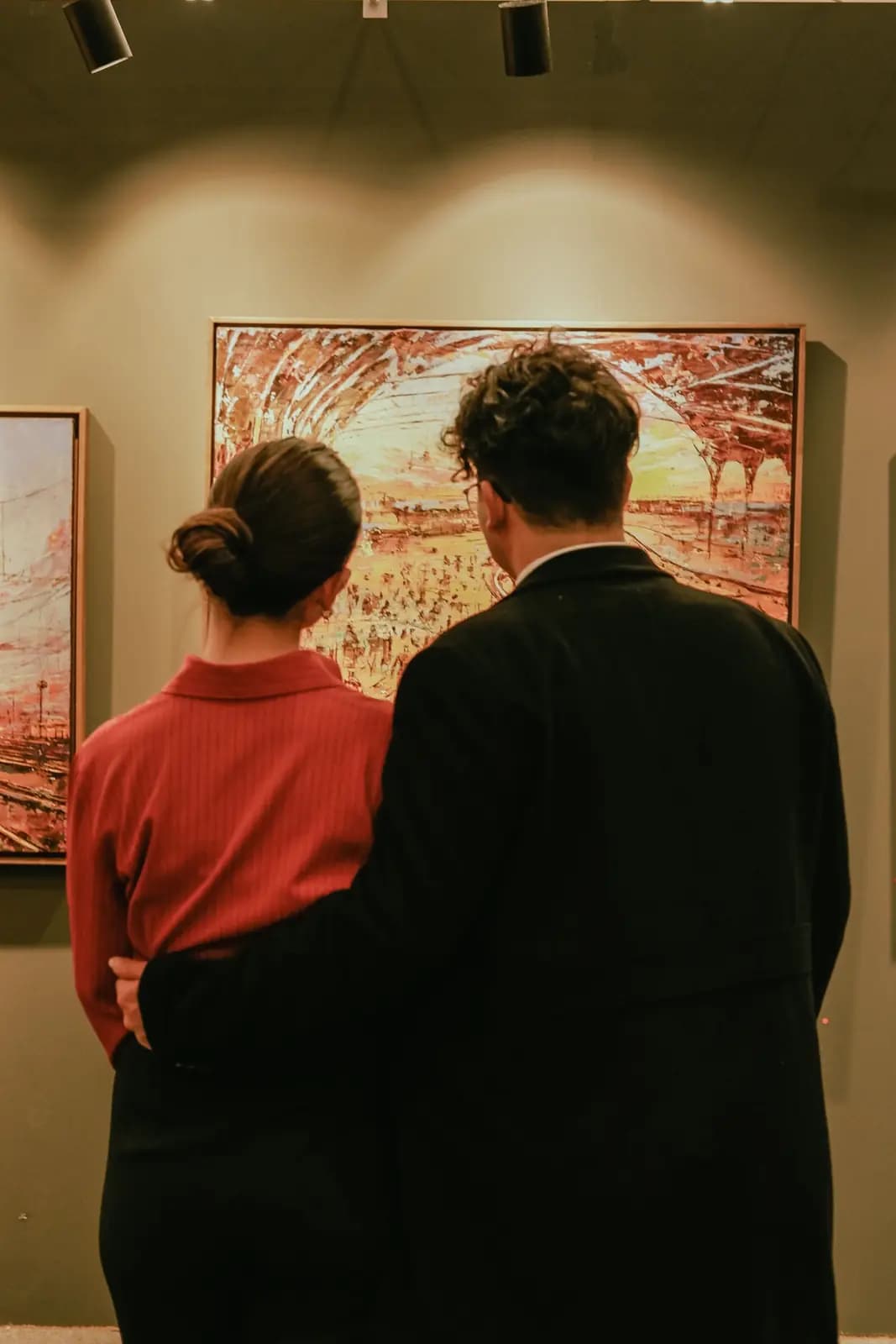 A couple stand arm in arm in front of a large painting under warm gallery lights, seen from behind.