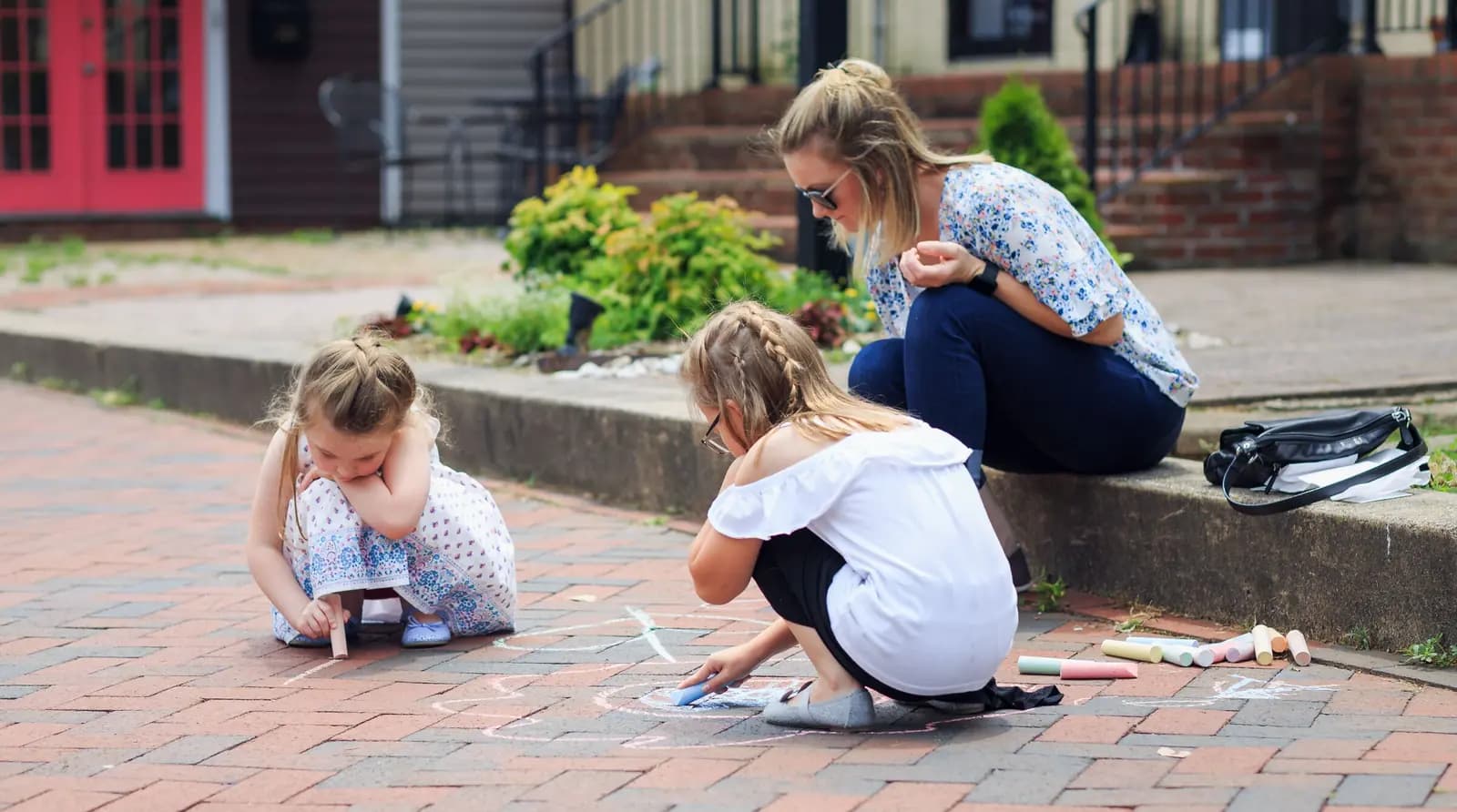 Two little girls crouch over chalk drawings on a brick sidewalk in front of houses while a parent sits on the curb beside them.