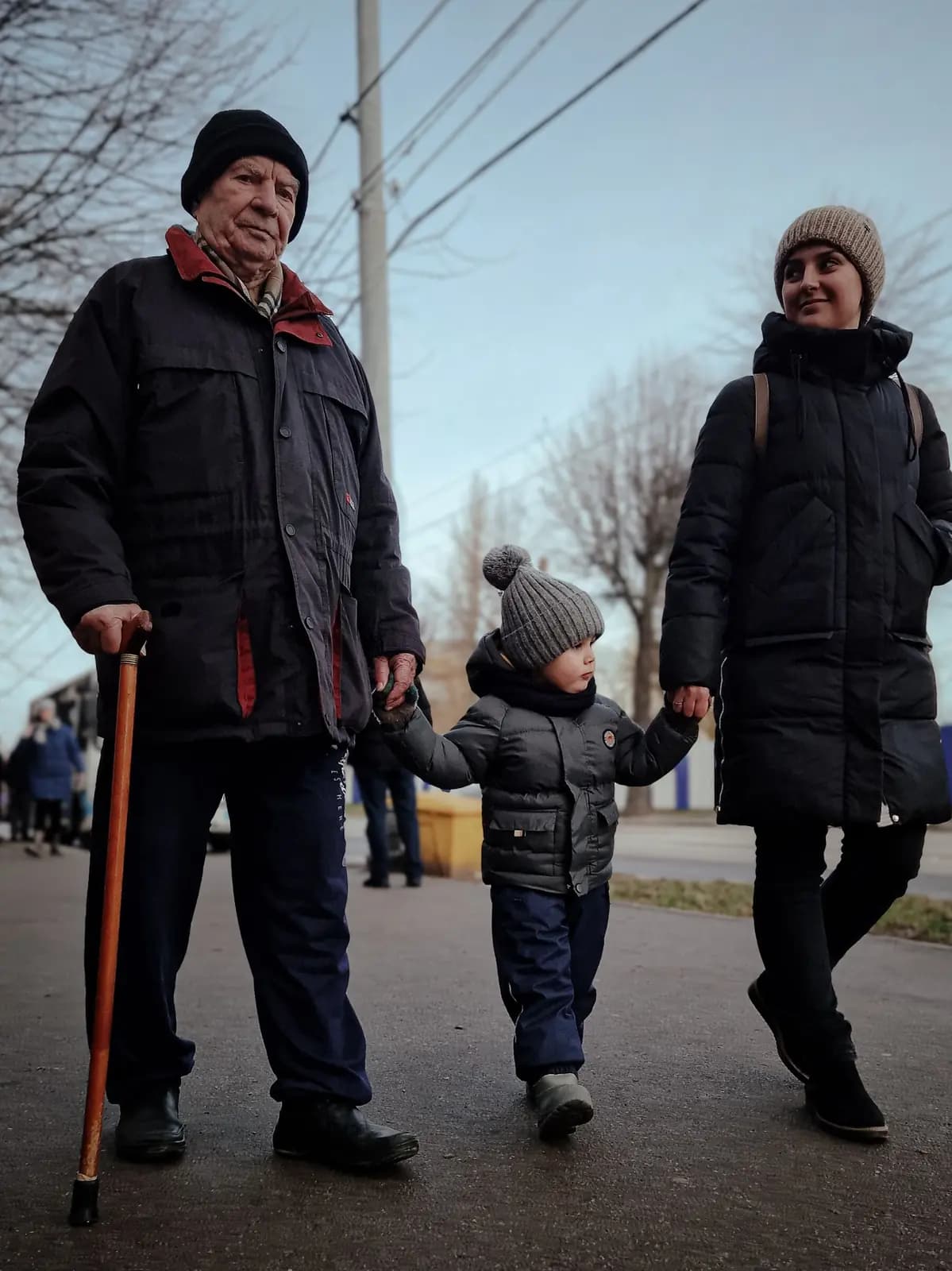 A grandfather with a cane and a parent walk hand in hand with a small boy between them on a sidewalk.