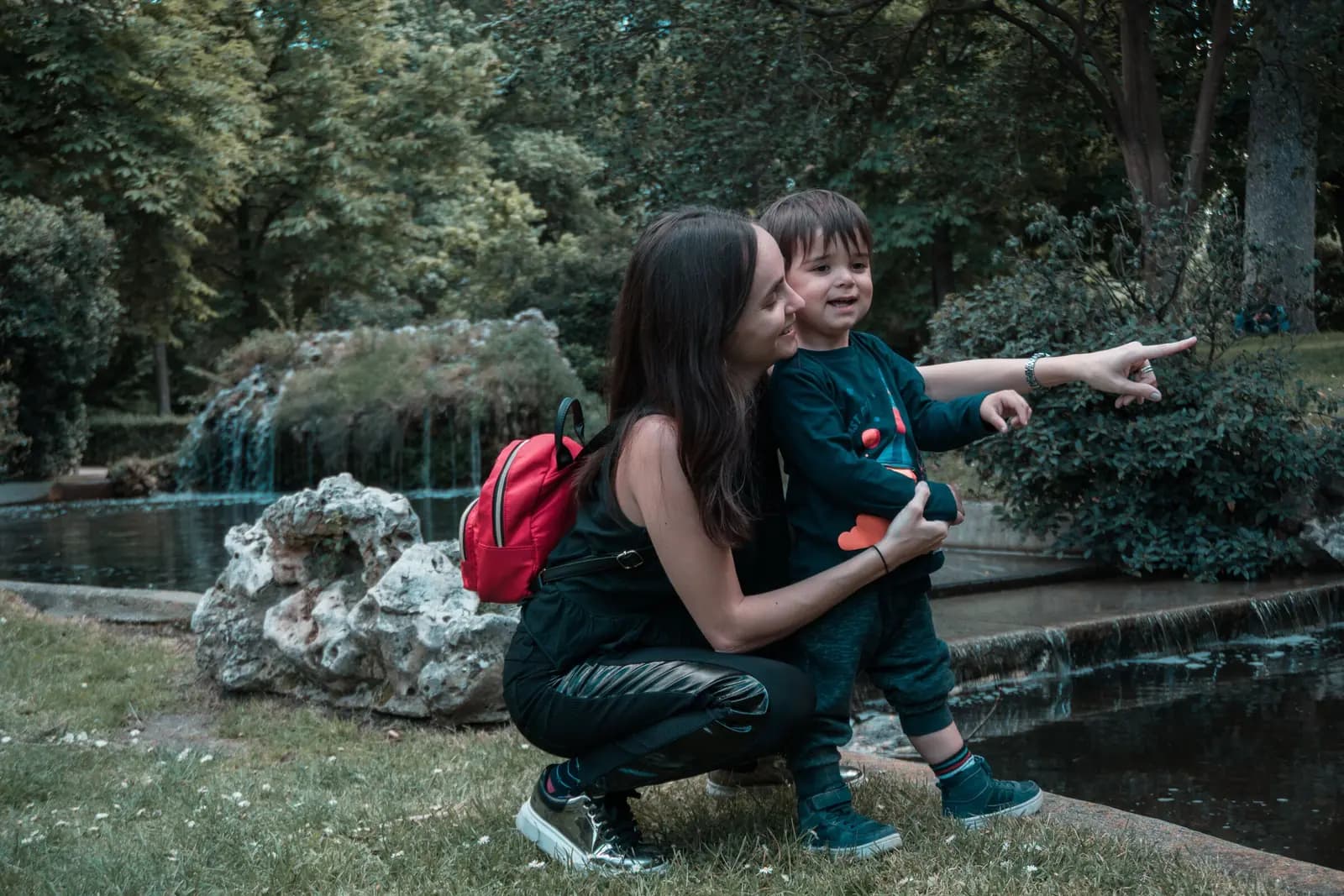 A parent crouches beside a toddler who points ahead, both looking the same way, in a park beside a pond.