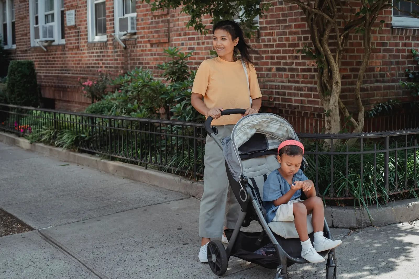 A parent pushes a stroller along a sidewalk past a brick building, looking across the street, with a small child in the seat.