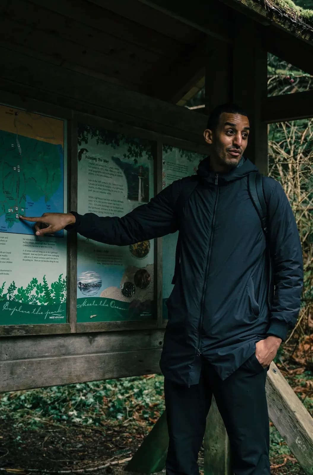 A man at a covered trailhead kiosk points at the trail map.