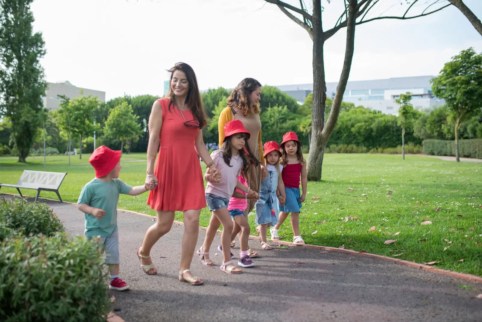 Two women walk along a park path with a group of children in bright red hats.