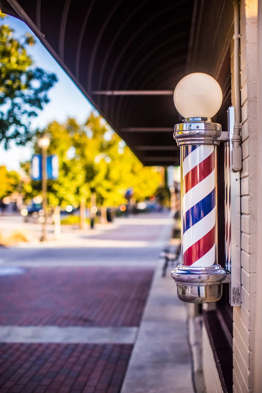 A lit barber pole on a brick shopfront beside a sunny small-town sidewalk.