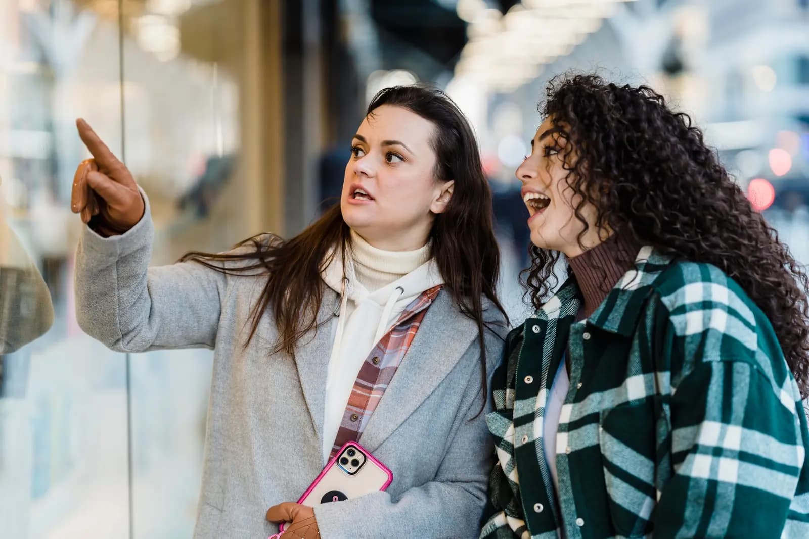 Two friends at a shop window, one pointing at the display while holding her phone.