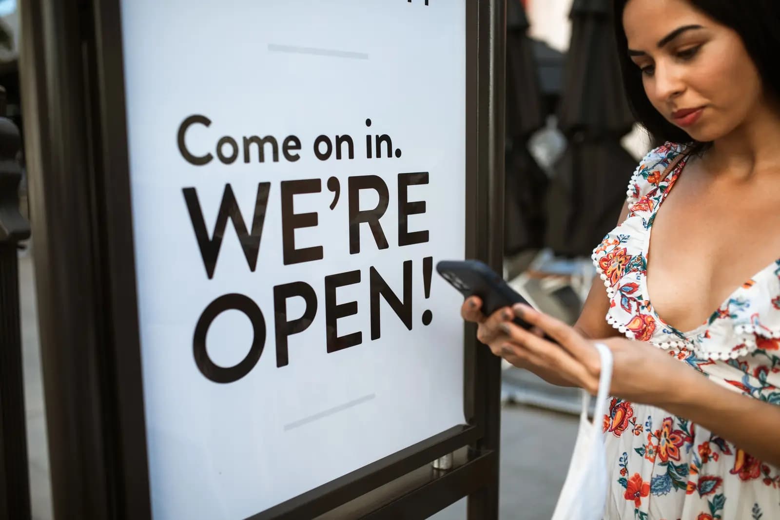 A woman checks her phone beside a large sign reading Come on in, we're open.