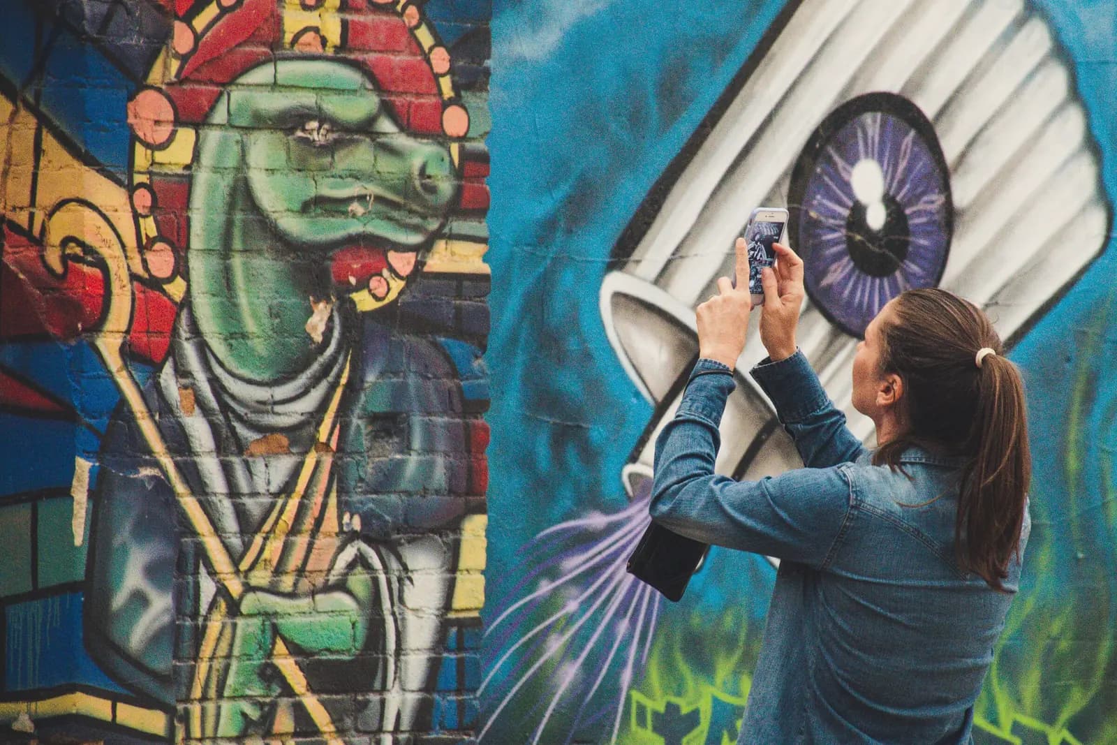 A woman holds up a phone to photograph a large painted mural on a brick wall.