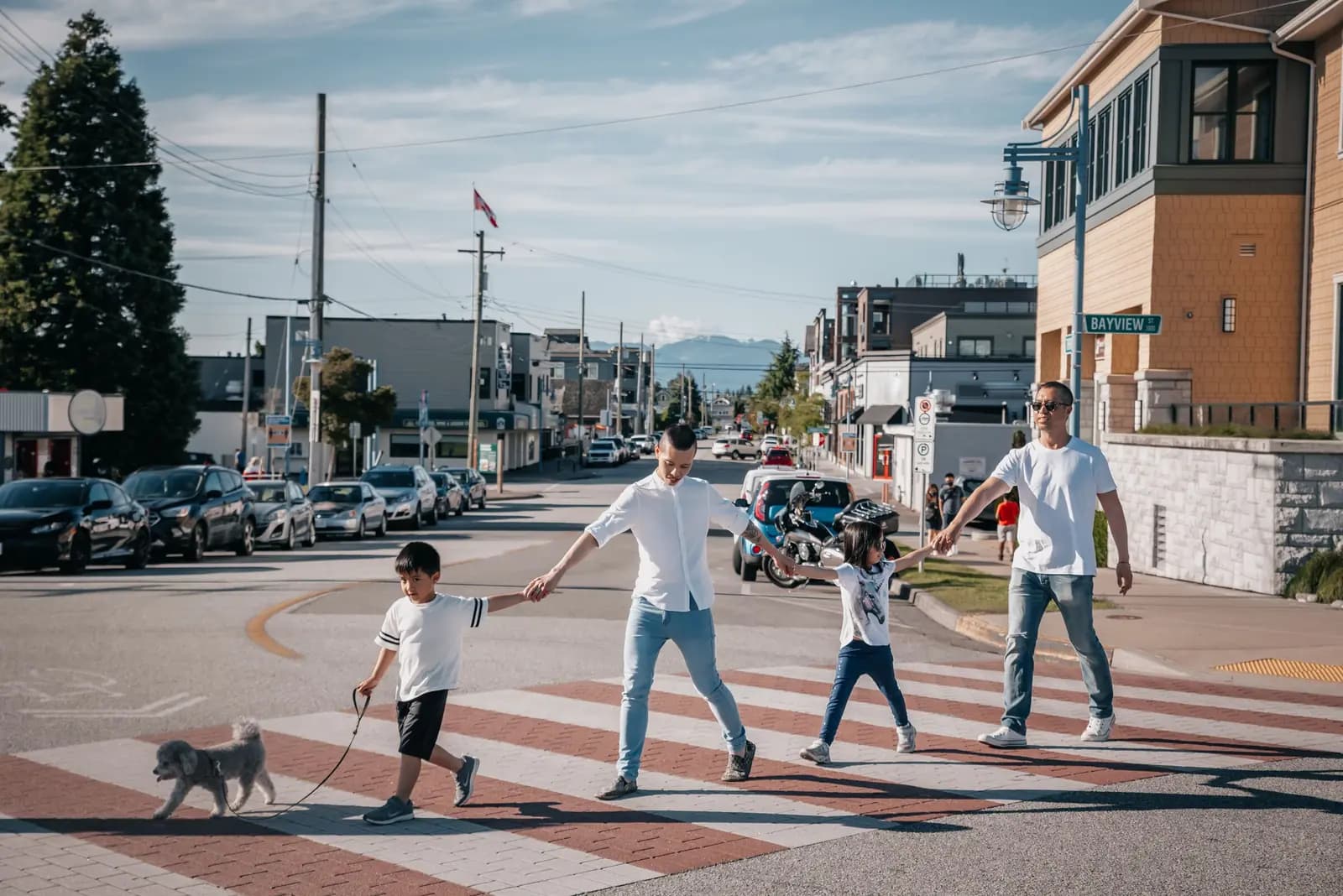Two parents and two children cross a sunny small-town street hand in hand with a little dog.