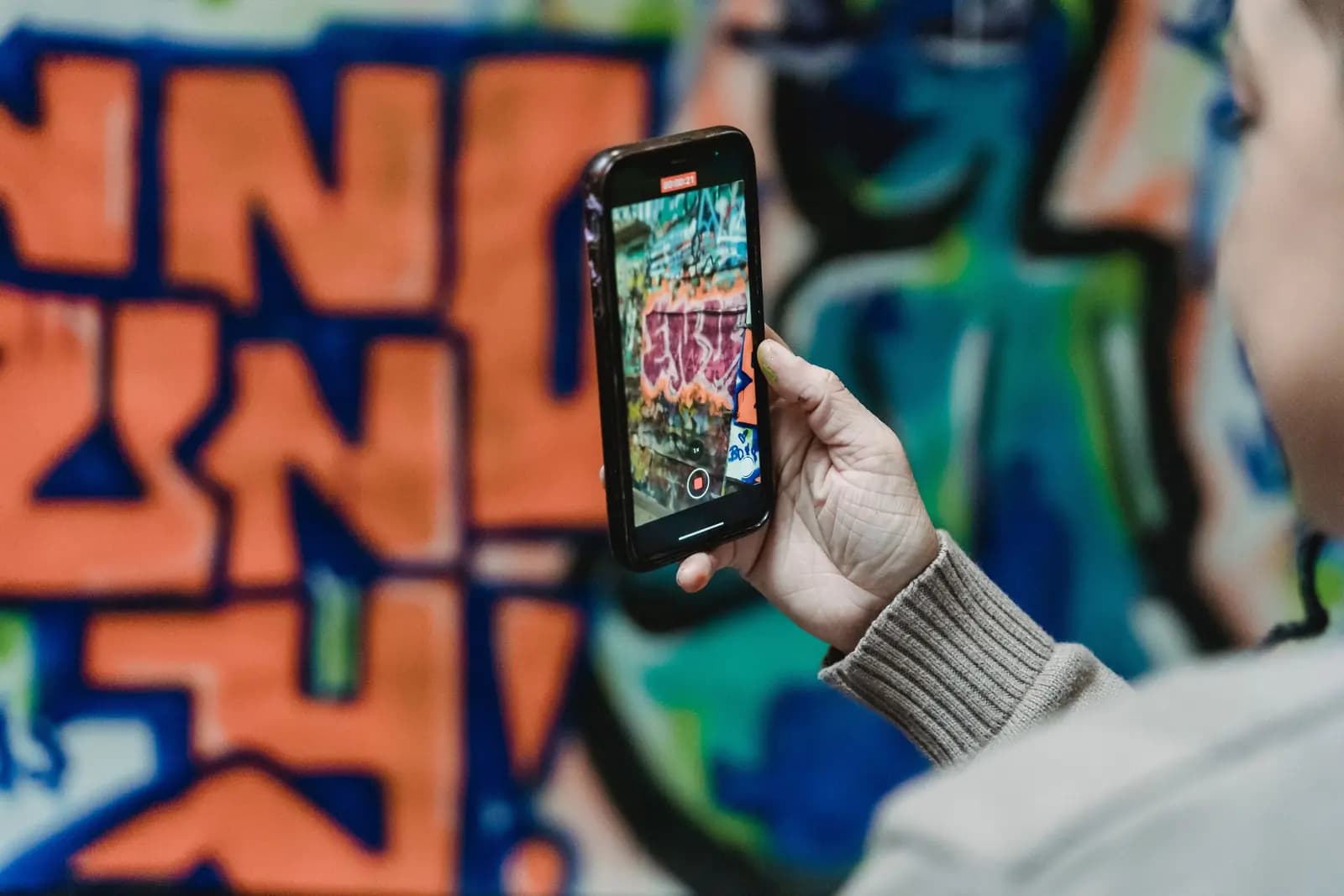 A hand holds up a phone to photograph a bright graffiti mural, with the mural on the screen.