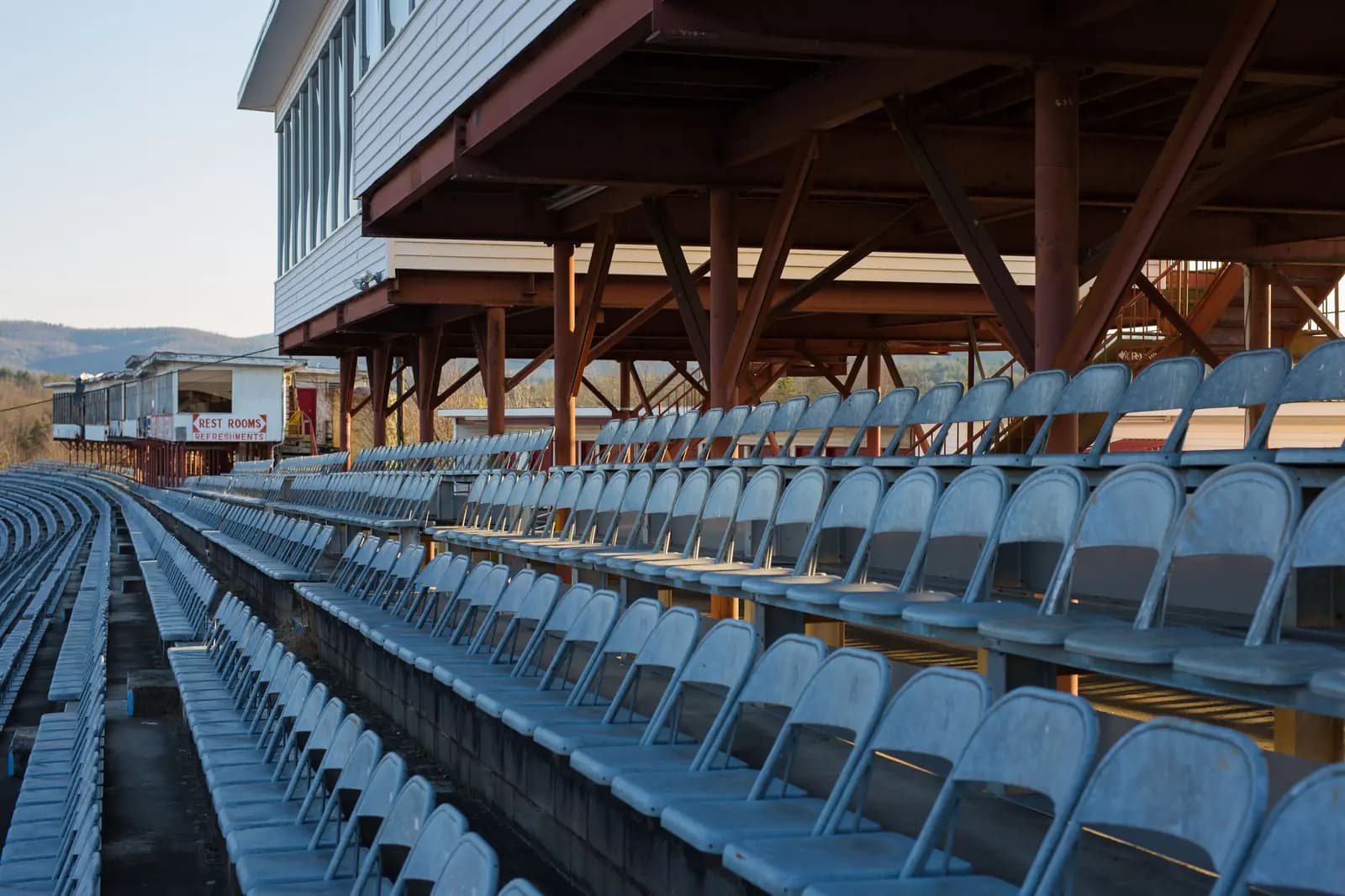 Rows of empty blue folding seats in a covered wooden grandstand at a small-town ballpark.