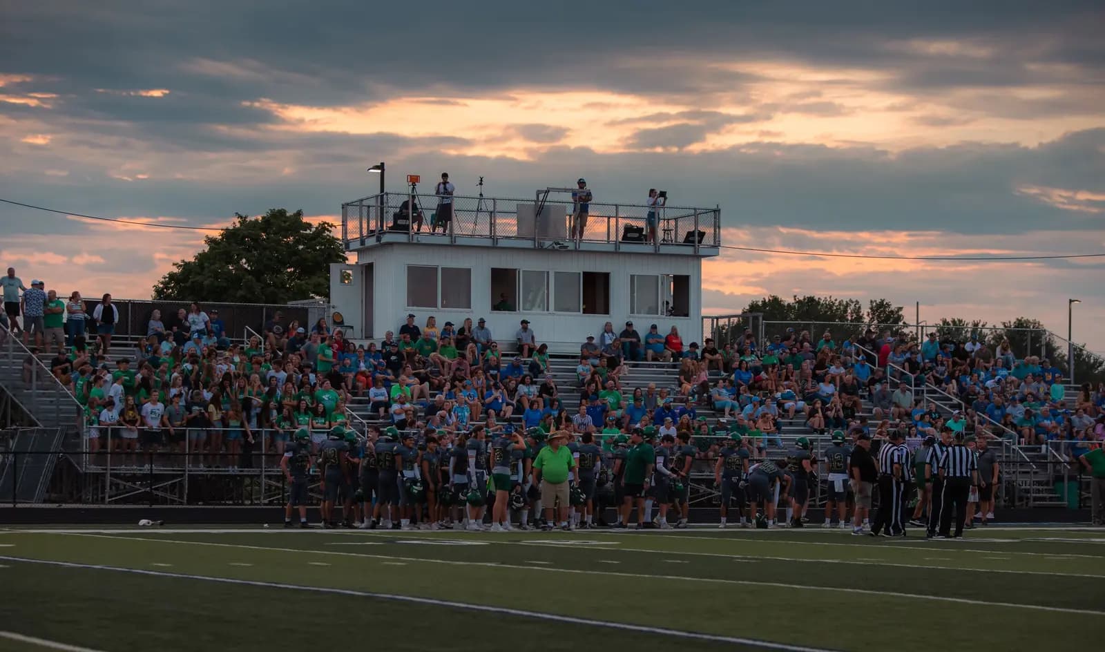A packed home grandstand and press box at a high school football game at dusk, with the team on the sideline.