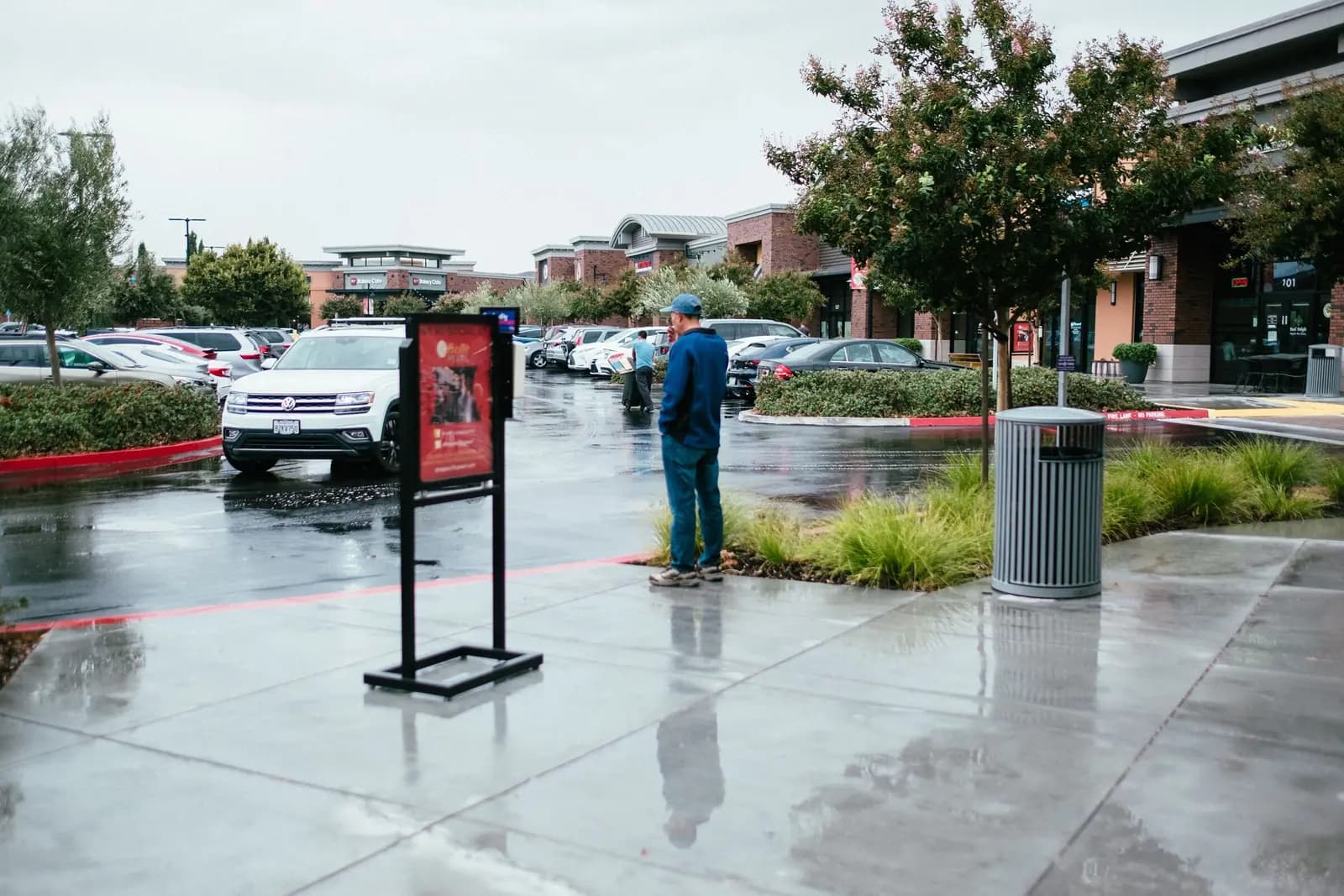 A man in a cap stands beside a sign on a stand in a wet shopping-center plaza.