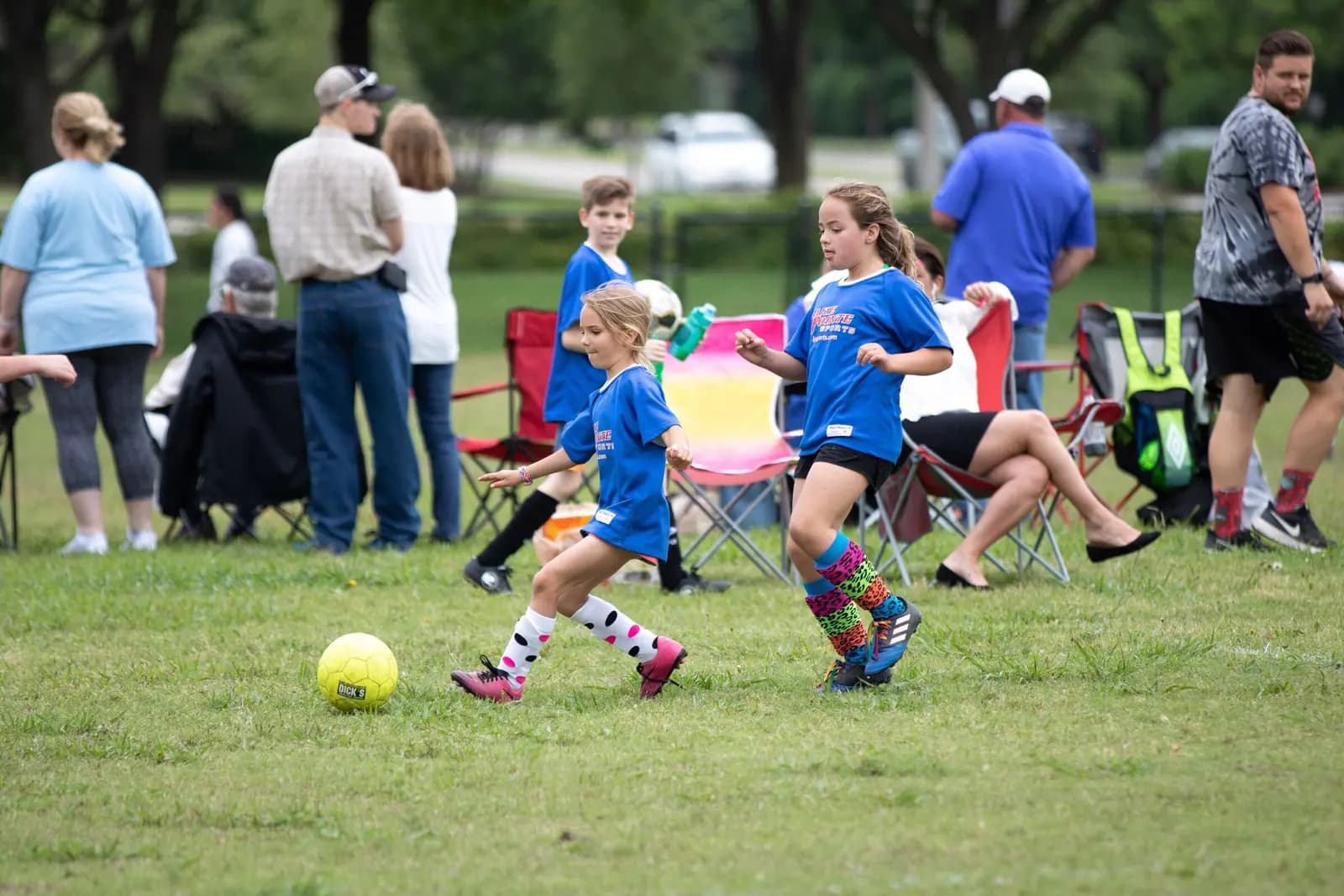 A girls' youth soccer game with parents in folding chairs and standing along the sideline behind the players.