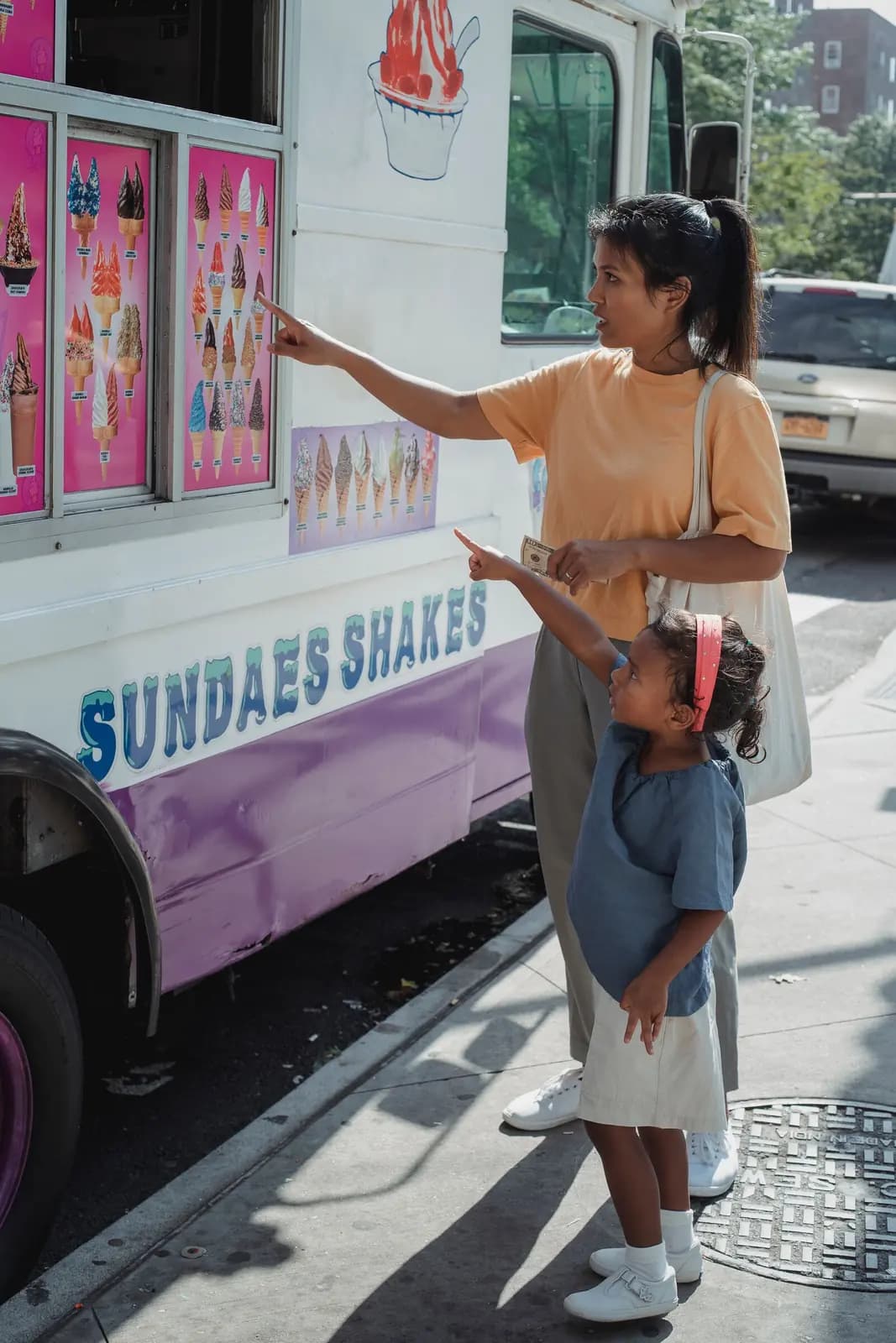 A mother and young daughter stand at an ice cream truck on a city sidewalk, the girl pointing up at the menu.