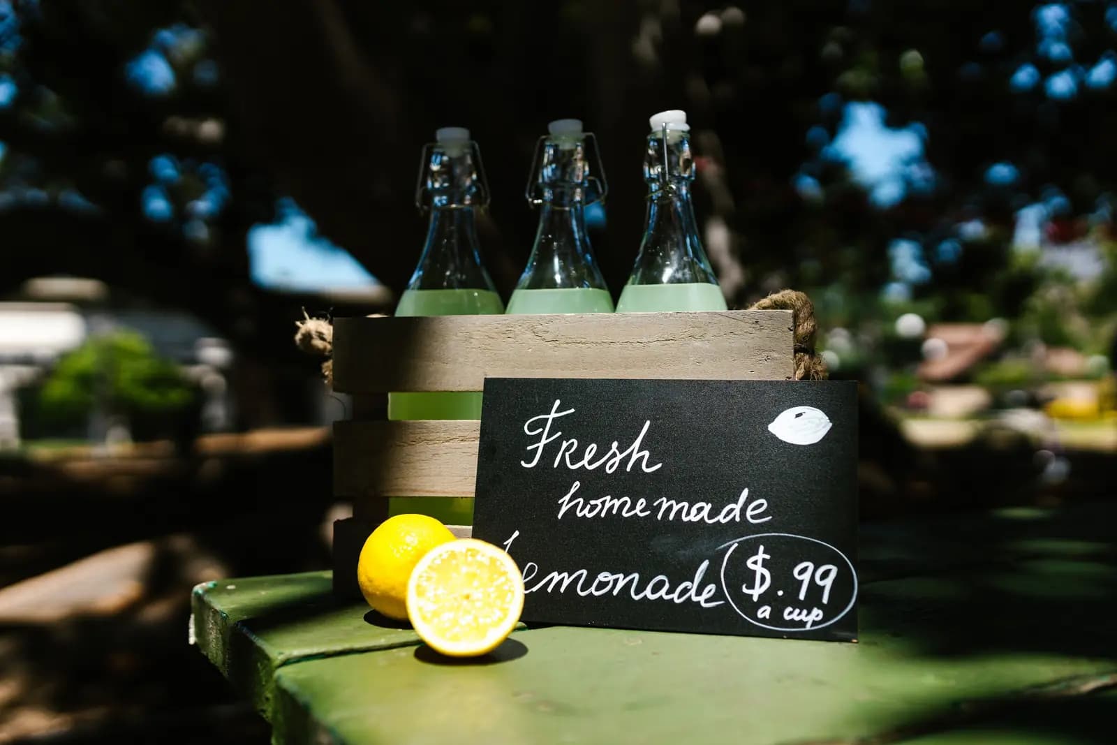A crate of bottled lemonade on a green table outdoors, with a chalkboard sign reading fresh homemade lemonade.