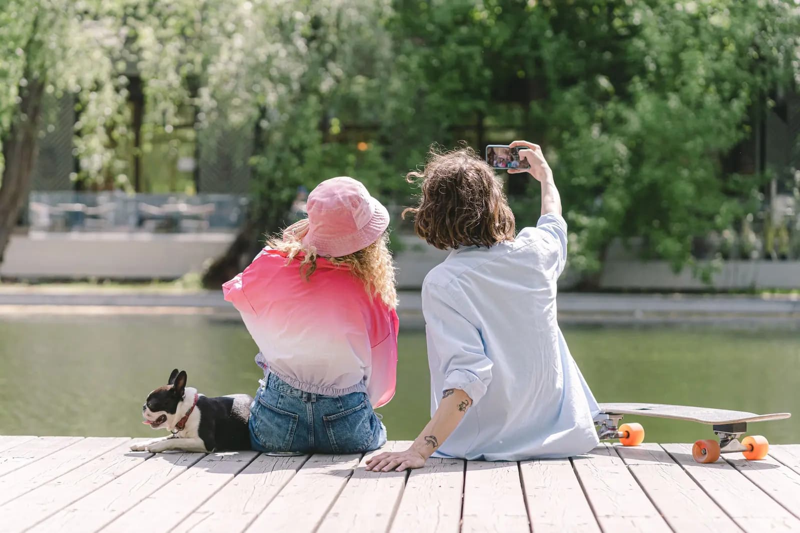 Two young people sit on a dock by the water with a dog and a skateboard, one holding a phone up to take a photo.