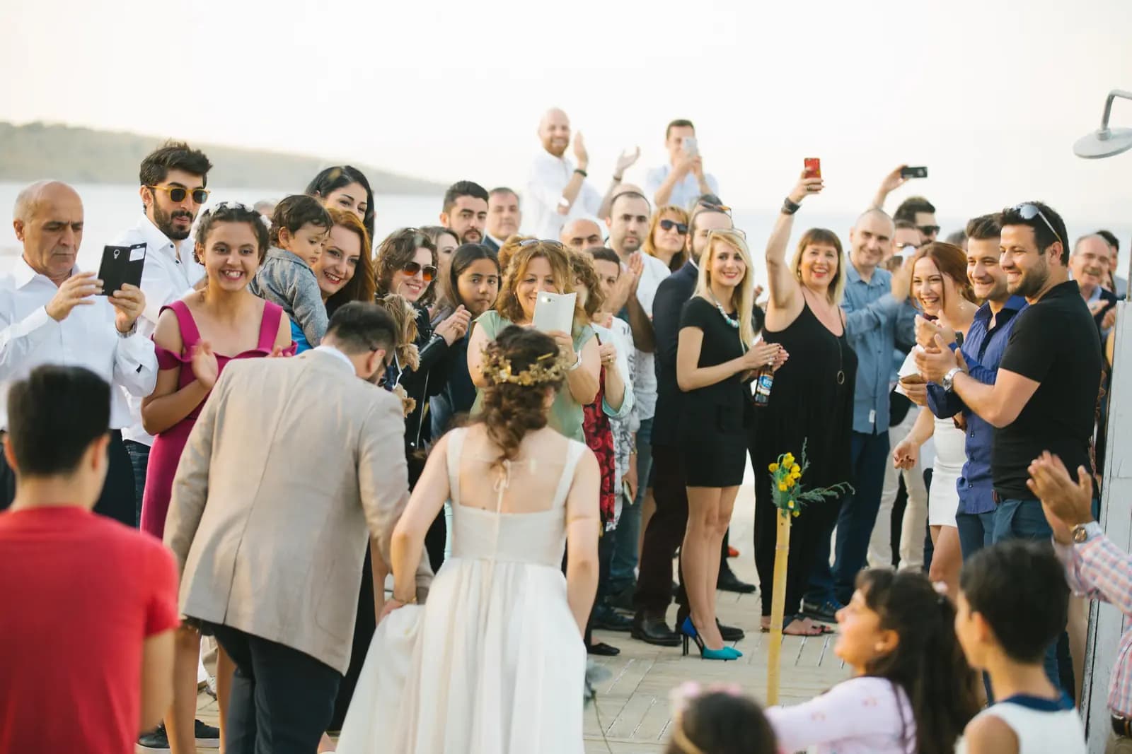 Wedding guests crowd together in daylight, many holding up phones and cheering as the bride arrives.
